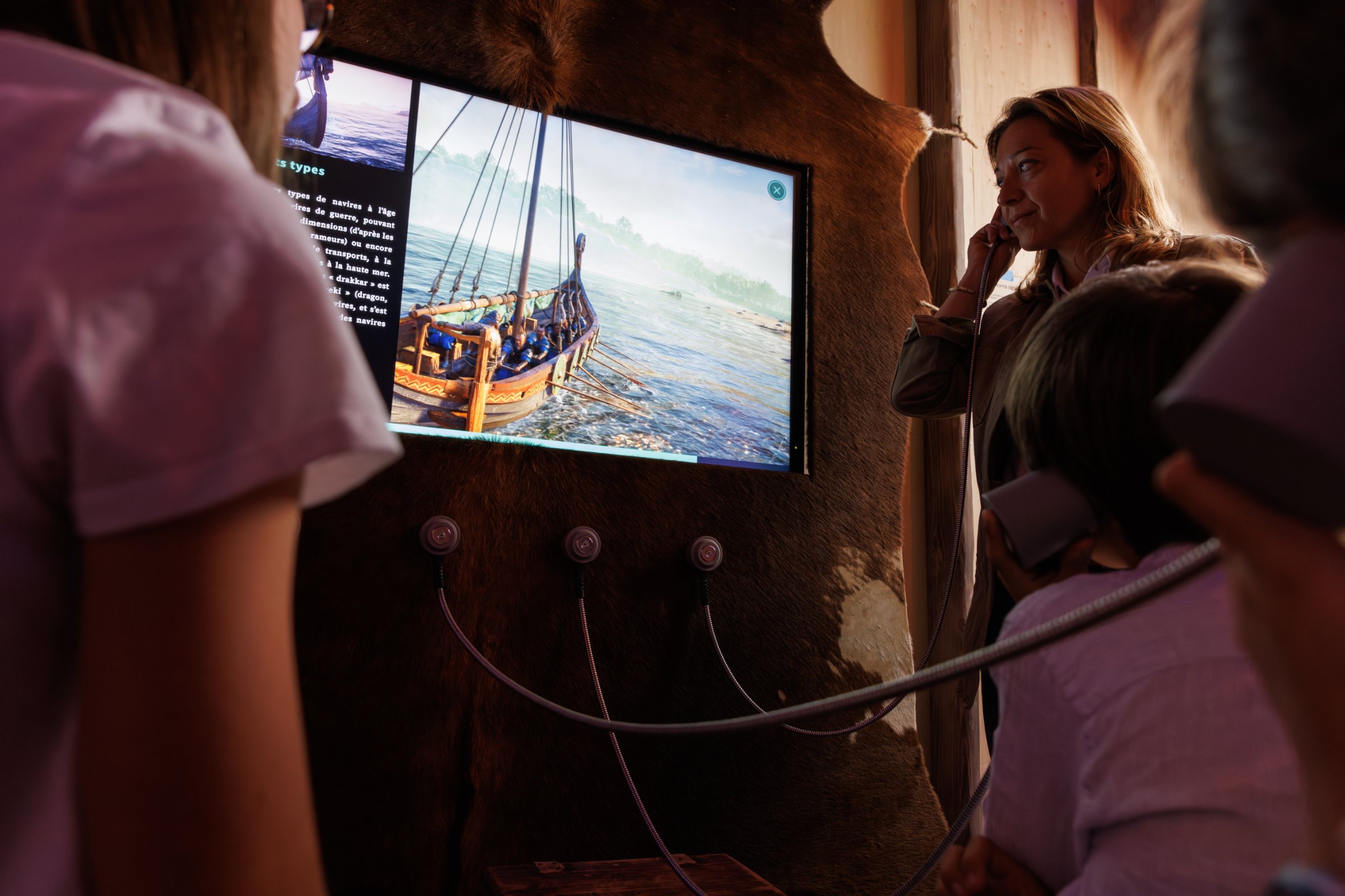 L'image montre un groupe de personnes, dont une femme et des enfants, observant un écran. L'écran présente une image d'un bateau sur l'eau, probablement dans un contexte historique ou éducatif. Les participants semblent écouter des informations à travers des casques audio reliés à l'écran. L'environnement est chaleureux, et les murs sont décorés de manière rustique, ce qui ajoute à l'atmosphère immersive de l'expérience.
