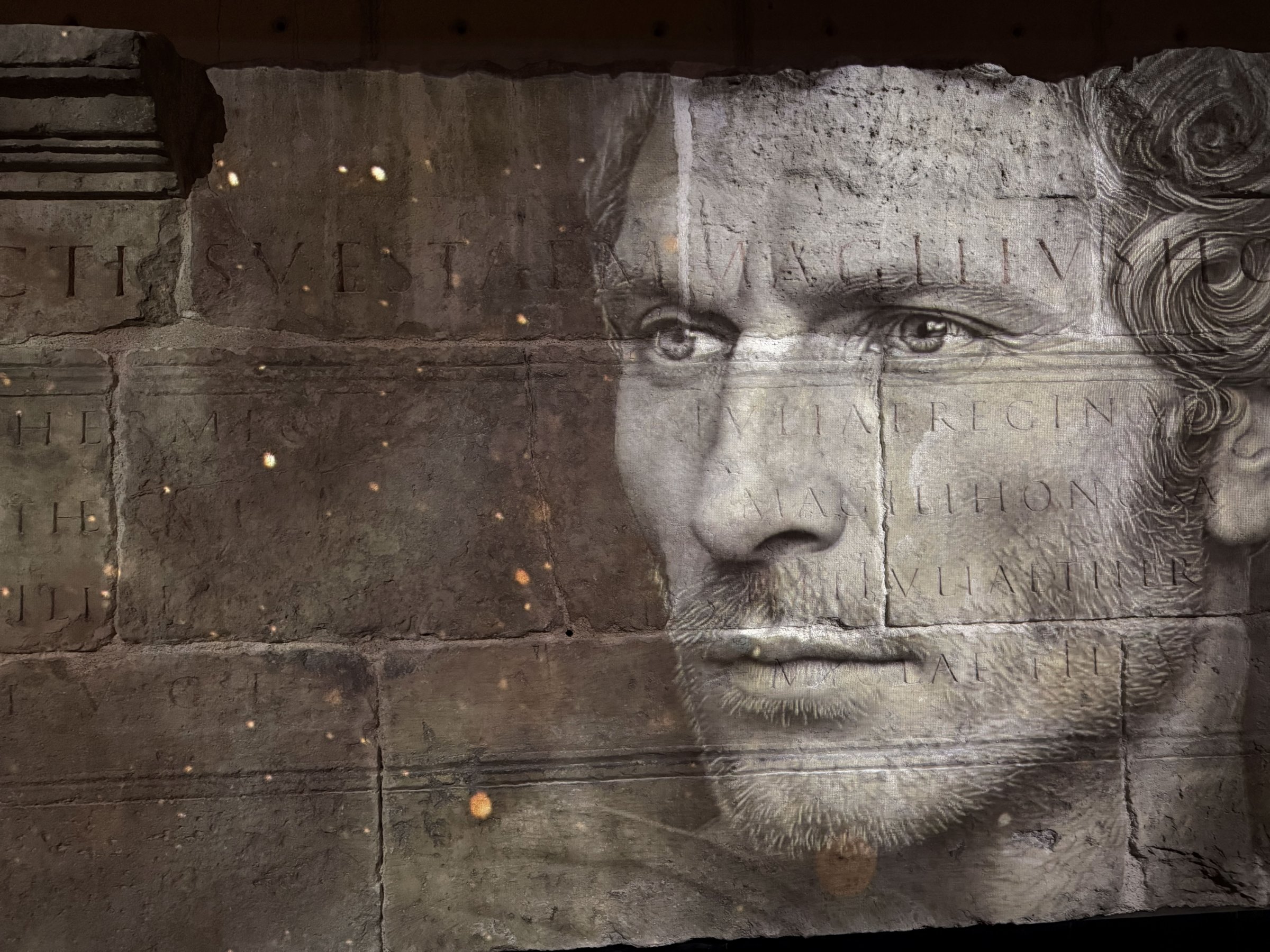 L'image présente une série de visages en noir et blanc, superposés de manière artistique sur une surface ressemblant à de la pierre ou du béton. Les traits des visages sont flous, créant une atmosphère mystérieuse. Au centre, des inscriptions en lettres capitales sont visibles, probablement des noms ou des titres. L'ensemble de la composition dégage une impression de gravité et d'histoire, suggérant une connexion avec des thèmes de mémoire ou d'honneur.