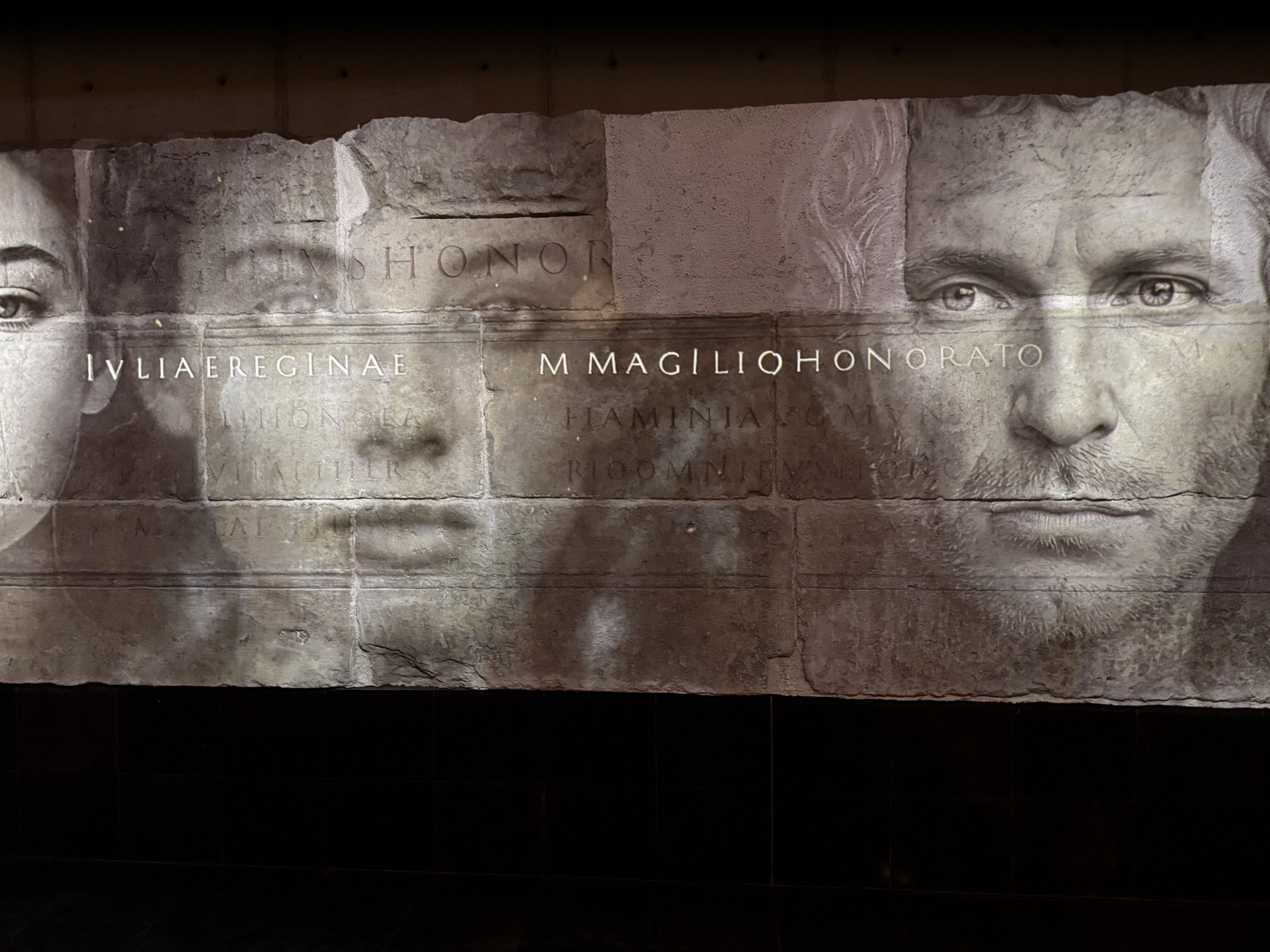 L'image présente une série de visages en noir et blanc, superposés de manière artistique sur une surface ressemblant à de la pierre ou du béton. Les traits des visages sont flous, créant une atmosphère mystérieuse. Au centre, des inscriptions en lettres capitales sont visibles, probablement des noms ou des titres. L'ensemble de la composition dégage une impression de gravité et d'histoire, suggérant une connexion avec des thèmes de mémoire ou d'honneur.