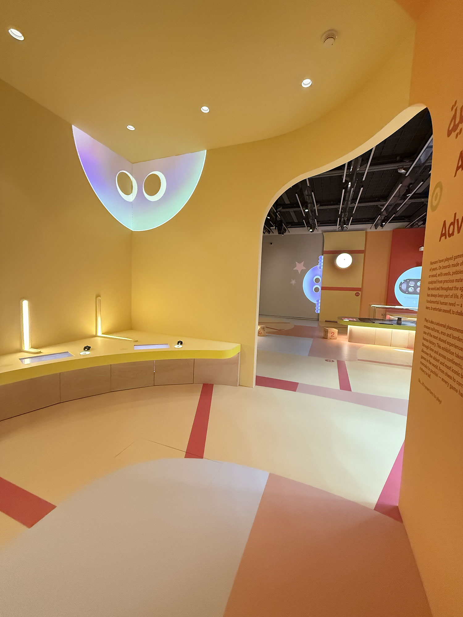L'image montre un espace intérieur aux murs de teintes jaunes, avec un éclairage doux et des éléments de design moderne. On y voit des courbes et des formes géométriques sur le sol, qui semblent être un mélange de couleurs pastel. Des affichages lumineux projettent des formes ou des visages stylisés sur les murs. On note aussi des zones de travail ou d'exploration, avec des tables et des sièges. L'ensemble crée une atmosphère accueillante et ludique.