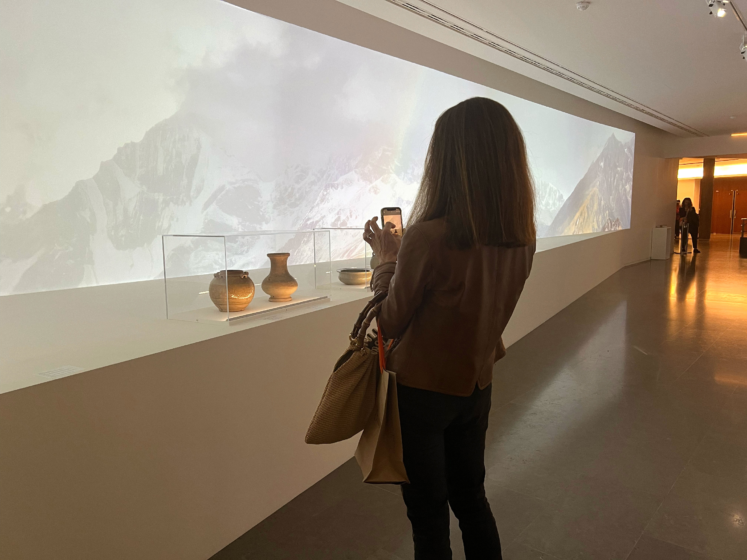 L'image montre une femme se tenant devant une exposition dans un musée. Elle est de dos, prenant une photo avec son téléphone. Devant elle, il y a une vitrine contenant plusieurs pots, tandis qu'en arrière-plan, on peut voir une projection de paysages de montagnes. L'éclairage est doux, et l'atmosphère semble calme et contemplative.