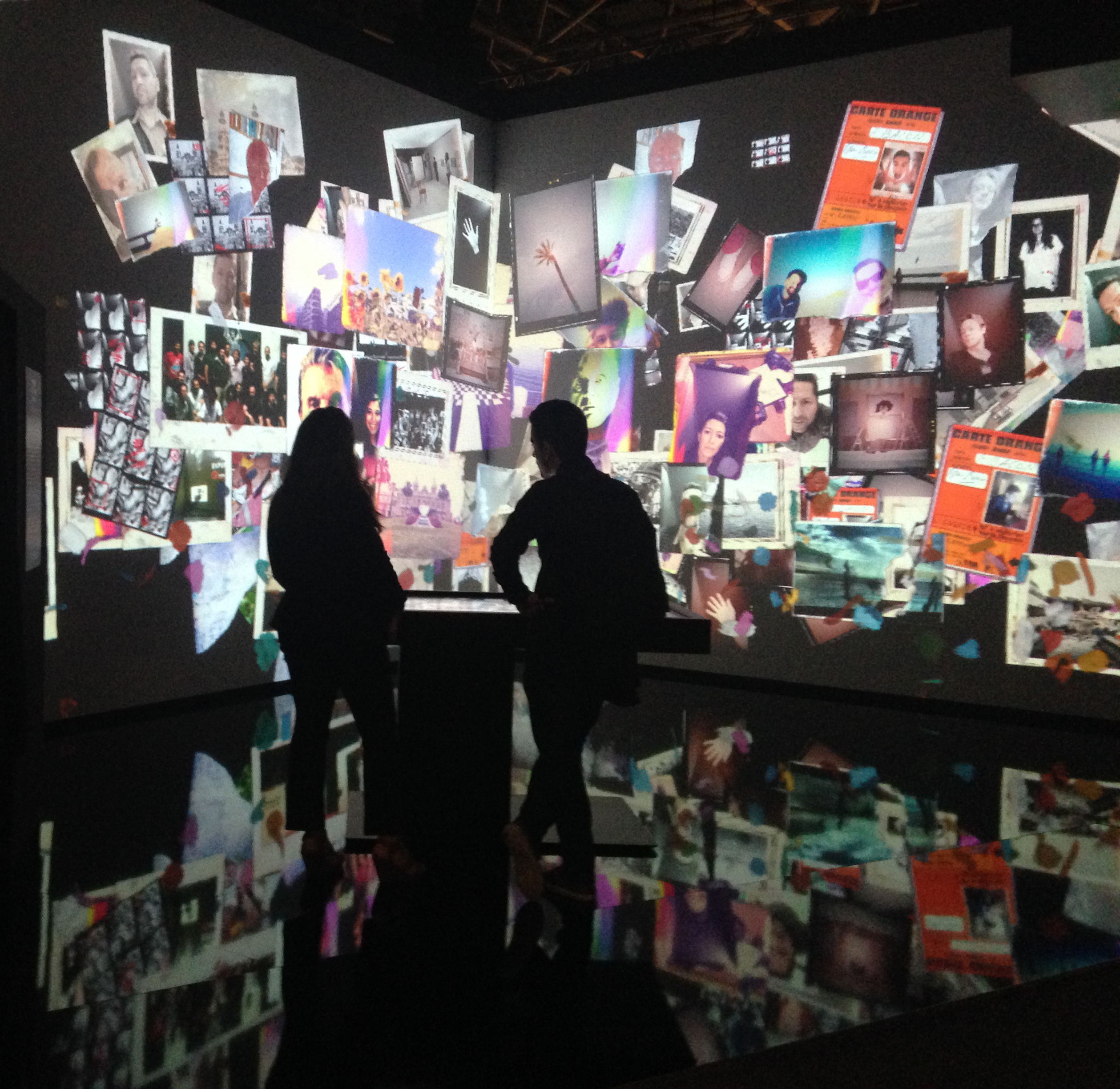 L'image montre une installation artistique immersive, où des projections de diverses photos et affiches couvrent les murs et le sol d'un espace sombre. Deux silhouettes de personnes se tiennent devant cette exposition, observant les images qui les entourent. L'ambiance semble à la fois moderne et artistique, avec une variété d'images qui évoquent différentes émotions et souvenirs.