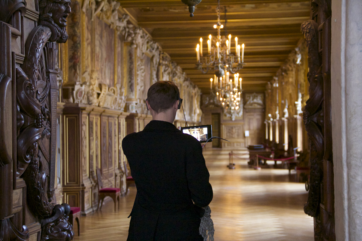 L'image montre une personne de dos, debout dans un long couloir d'un palais ou d'un manoir. Le couloir est orné de décorations élaborées et de sculptures en bois. De grands chandeliers pendent du plafond, illuminant l'espace avec une lumière chaleureuse. Des tableaux et des tapisseries ornent les murs, ajoutant à l'atmosphère historique. La personne tient un appareil photo ou une tablette, semblant capturer la beauté de l'architecture environnante.