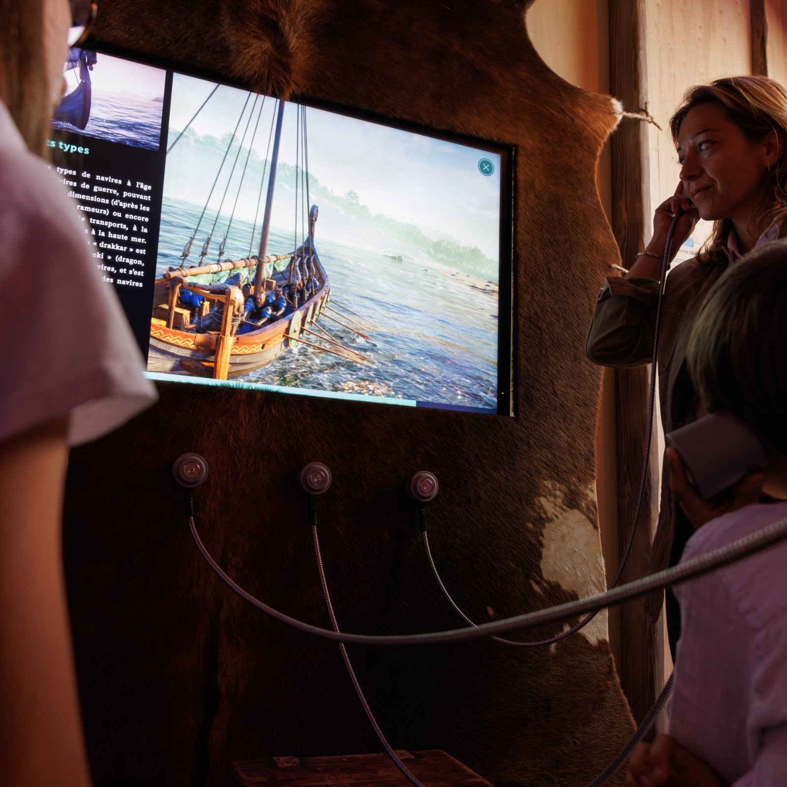 L'image montre un groupe de personnes, dont une femme et des enfants, observant un écran. L'écran présente une image d'un bateau sur l'eau, probablement dans un contexte historique ou éducatif. Les participants semblent écouter des informations à travers des casques audio reliés à l'écran. L'environnement est chaleureux, et les murs sont décorés de manière rustique, ce qui ajoute à l'atmosphère immersive de l'expérience.