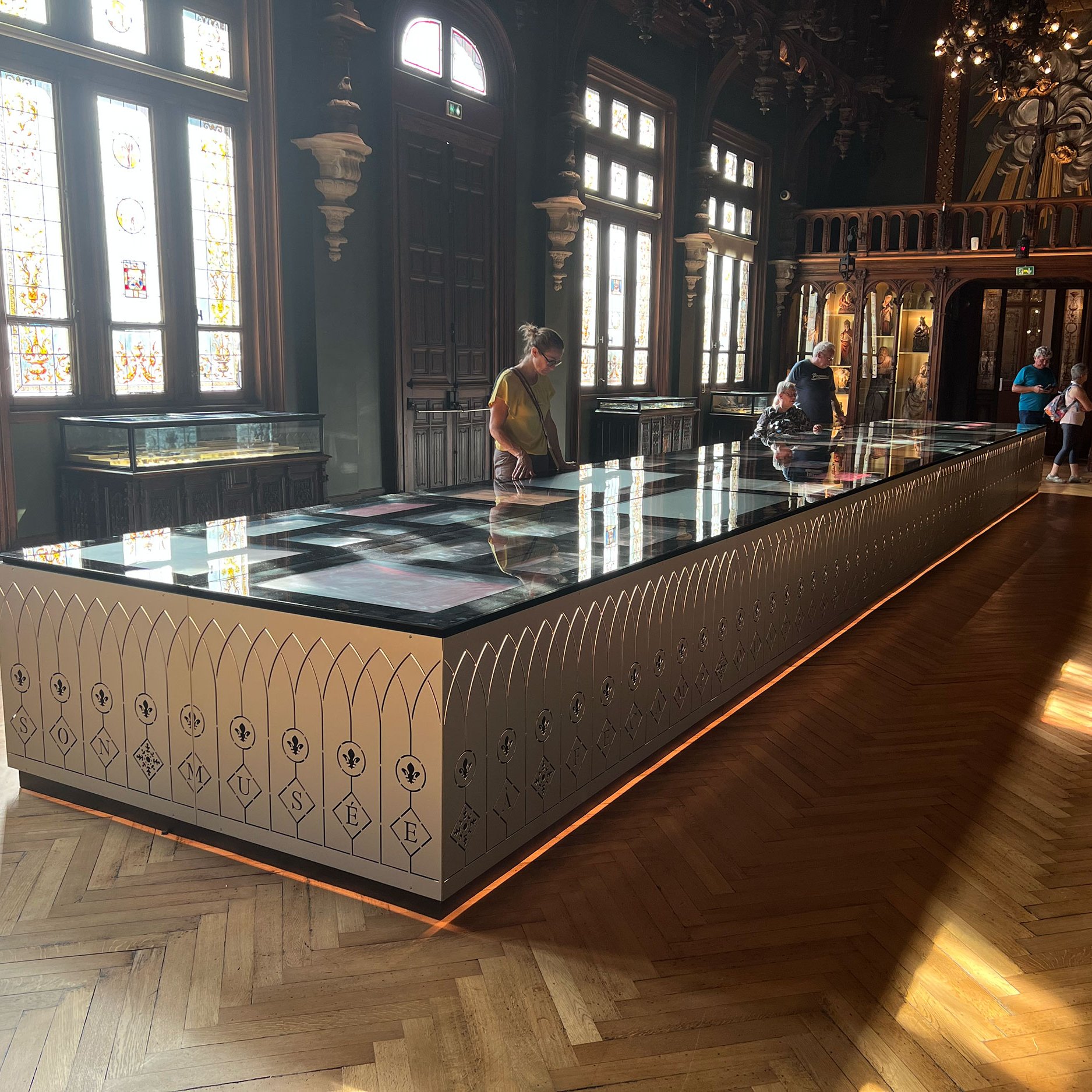 L'image montre une salle intérieure d'un musée ou d'un palais, avec un éclairage naturel provenant des fenêtres. Au centre, il y a une grande vitrine rectangulaire en verre qui semble exposer des objets, peut-être des artefacts ou des œuvres d'art. Le mobilier présente un design élégant avec des motifs décoratifs sur les côtés. Le sol est en bois avec un motif de parquet en chevron. Des visiteurs inspectent la vitrine, offrant une idée de l'ambiance paisible et muséale du lieu. Les murs sont ornés de détails architecturaux raffinés.