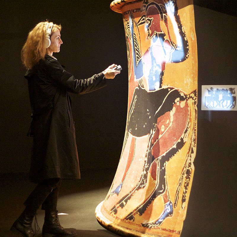 L'image montre une femme en train d'interagir avec un objet d'art, probablement une grande jarre ou un vase antique. Celui-ci est décoré de peintures représentant des figures humaines ou des scènes. La femme, vêtue d'un manteau noir, semble fasciné par la représentation sur le vase, ce qui crée une ambiance d'exploration et d'appréciation de l'art historique dans un cadre muséal ou d'exposition. L'éclairage met en valeur les détails des peintures, soulignant leur importance.