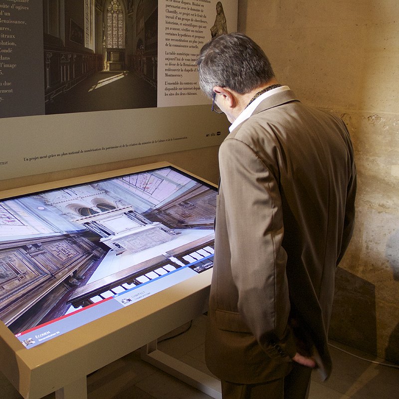 L'image montre un homme debout devant un grand écran tactile dans un espace intérieur. L'écran affiche une vue en 3D ou une reconstitution numérique d'une salle, probablement d'une église ou d'un lieu historique, mettant en évidence une architecture détaillée. L'environnement autour de l'écran est décoré de manière sobre, avec des murs en pierre et des éléments d'information visibles. L'homme semble attentif et intéressé par ce qu'il découvre à l'écran.
