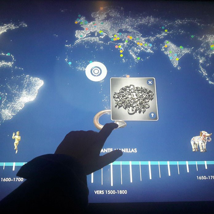 L'image semble représenter une interface interactive montrant une carte du monde avec des points lumineux indiquant différentes localisations. En bas, il y a une frise chronologique qui s'étend de 1500 à 1800, avec des étiquettes pour différentes périodes. Au centre, un élément avec des motifs abstraits est sélectionné, et une main de personne interagissant avec l'écran est visible. Des icônes ou illustrations, comme une statue et un éléphant, apparaissent également, ce qui suggère une exploration de l'histoire ou de la culture.
