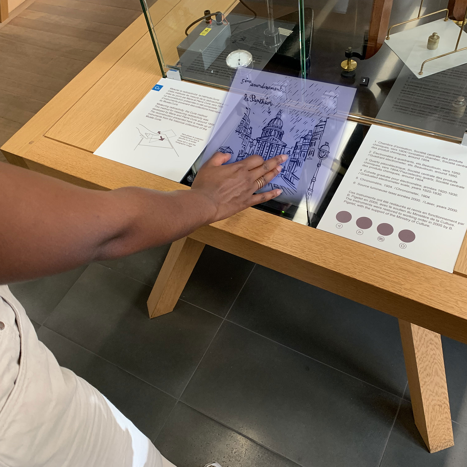 L'image montre une personne en train d'interagir avec un écran tactile placé sur une table en bois. On peut voir une représentation graphique d'un bâtiment, probablement une illustration d'un monument ou d'un lieu historique. À côté de l'écran, il y a des panneaux explicatifs contenant des informations. L'espace semble être une exposition, peut-être dans un musée ou une galerie. L'ensemble de la scène dégage une ambiance éducative et interactive.