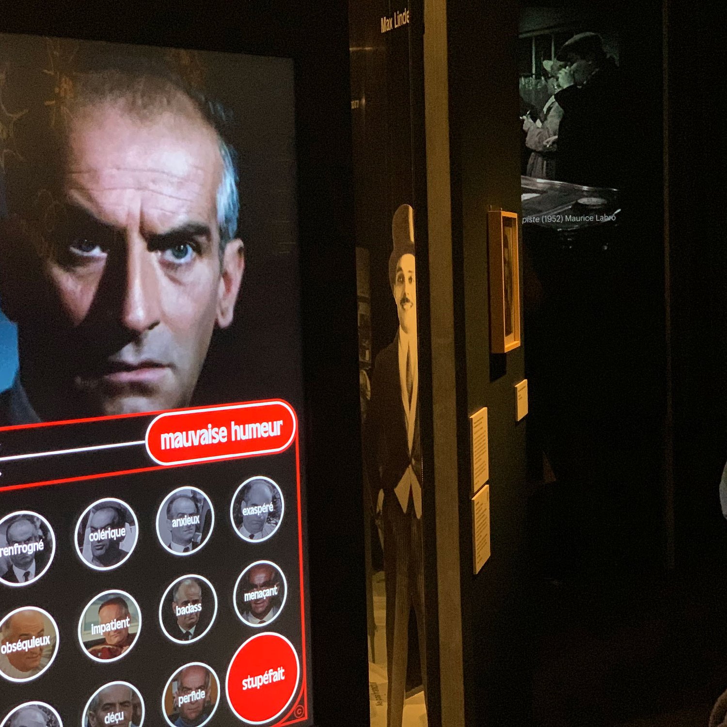 L'image montre une installation muséale avec un homme dont le visage est affiché sur un écran. Il regarde l'objectif avec une expression sérieuse. En bas de l'écran, il y a des mots qui semblent décrire différentes humeurs. Une personne portant un masque observe l'écran, suggérant une interaction avec l'affichage. À l'arrière-plan, on peut voir d'autres images en noir et blanc, ajoutant une atmosphère historique à l'ensemble de l'installation.