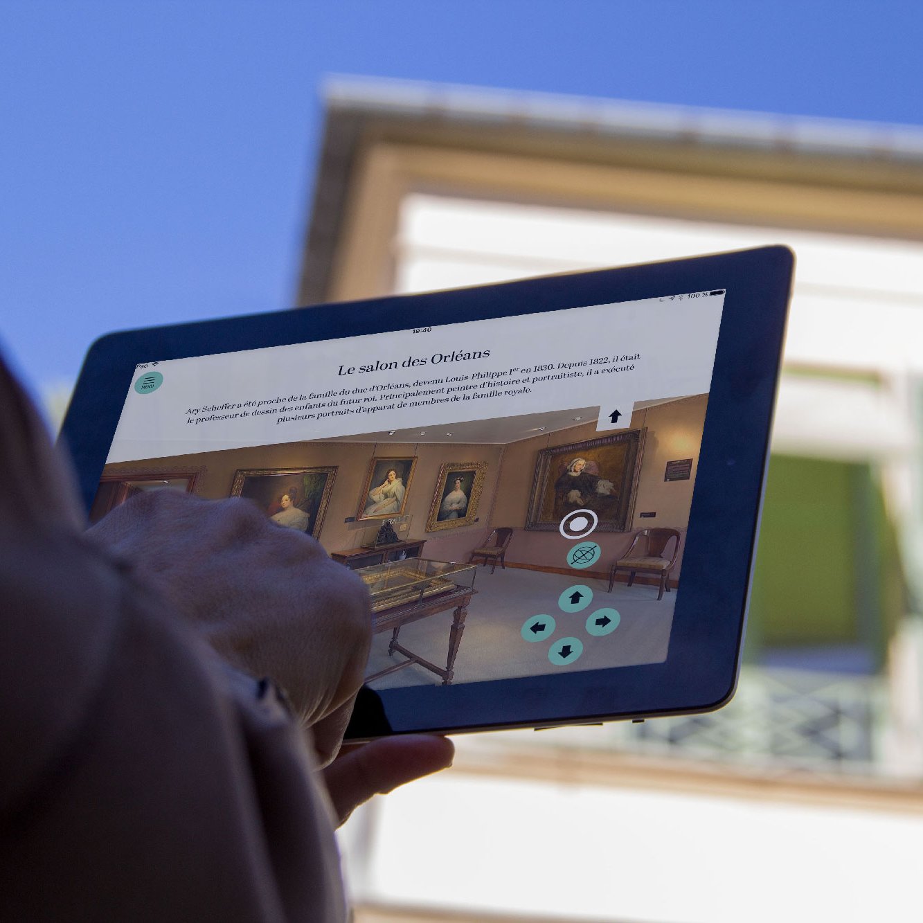 L'image montre une personne tenant une tablette dans un environnement extérieur, probablement près d'un musée ou d'une galerie d'art. Sur l'écran de la tablette, on peut voir une interface numérique qui semble présenter des informations sur une exposition, intitulée "Le salon des Oeuvres". En arrière-plan, il y a un bâtiment avec des volets ouverts et un ciel bleu, créant une ambiance agréable.