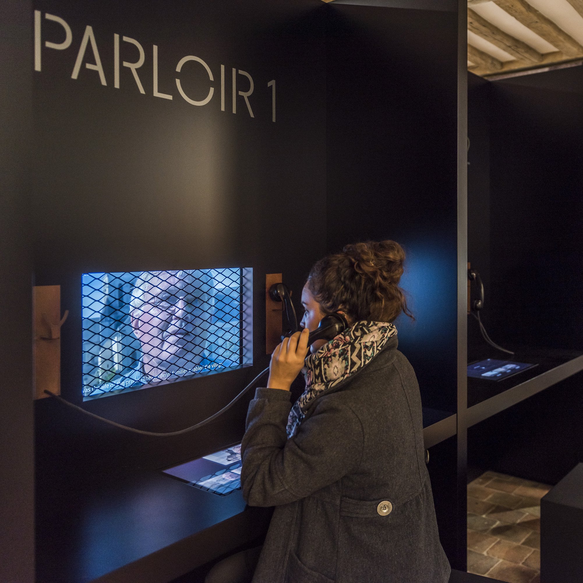 L'image montre un espace intérieur avec des cabines ou des sections séparées par des panneaux noirs. Une femme est assise à une table, utilisant un téléphone. Elle porte un manteau et une écharpe. À l'arrière-plan, on aperçoit des chaises alignées dans une pièce qui semble être un ancien lieu public ou historique. Un écran est visible, montrant une image à travers une grille. L'ambiance générale suggère un lieu de dialogue ou de communication, peut-être dans un cadre muséal ou éducatif.