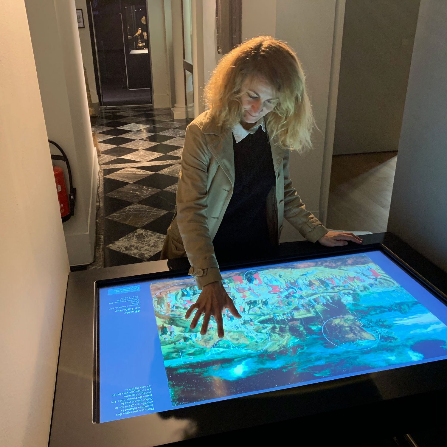 Sur l'image, on voit une femme aux cheveux blonds bouclés qui interagit avec un écran tactile. Elle semble examinée une œuvre d'art ou une illustration colorée affichée sur la surface de la table. L'environnement est élégant, avec un sol en damier noir et blanc, et on aperçoit des murs clairs. Dans le fond, des portes et des couloirs suggèrent un espace muséal. La scène dégage une atmosphère de curiosité et d'exploration.