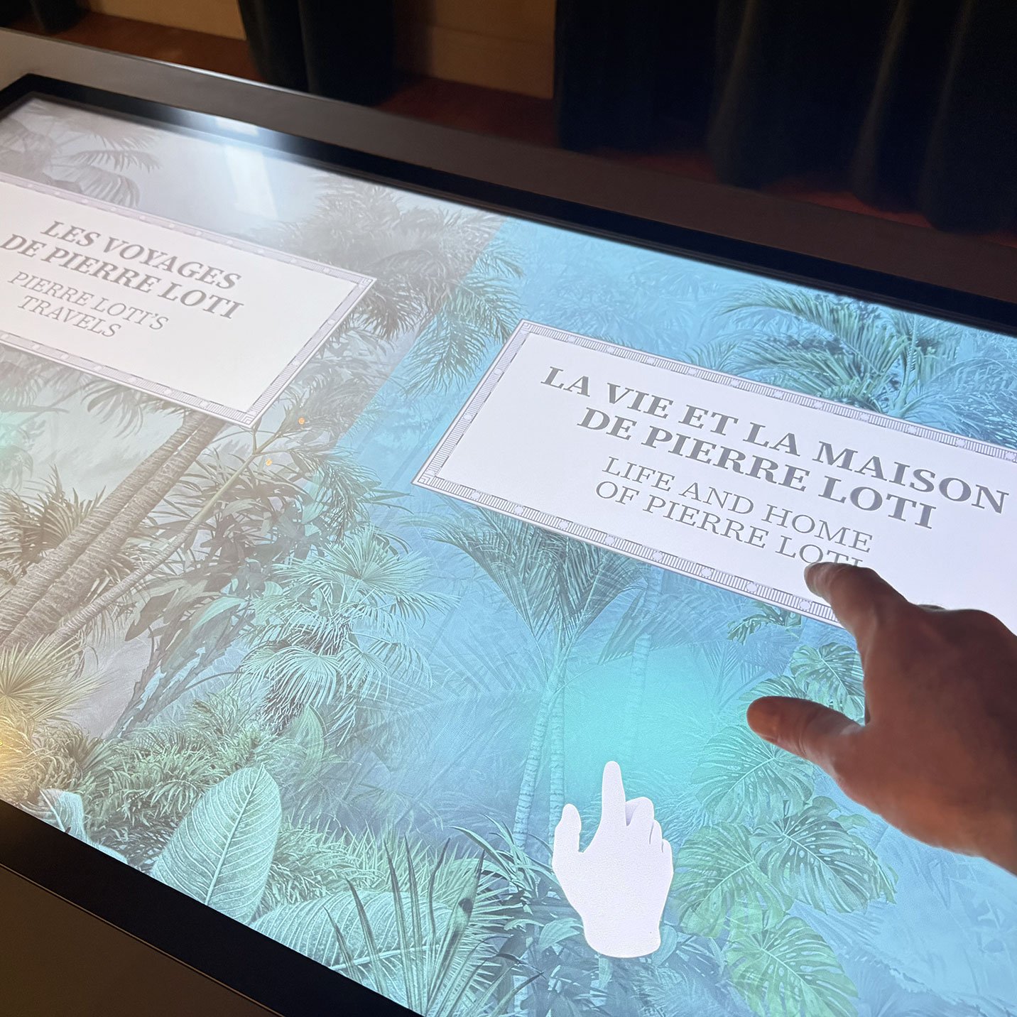 L'image montre un écran tactile interactif avec deux sections principales. À gauche, on peut lire "Les voyages de Pierre Loti", et à droite, "La vie et la maison de Pierre Loti", en français et en anglais. En bas, une main pointe vers l'écran, suggérant une interaction. L'arrière-plan est une illustration de paysages tropicaux, évoquant probablement les thèmes des voyages de Pierre Loti.