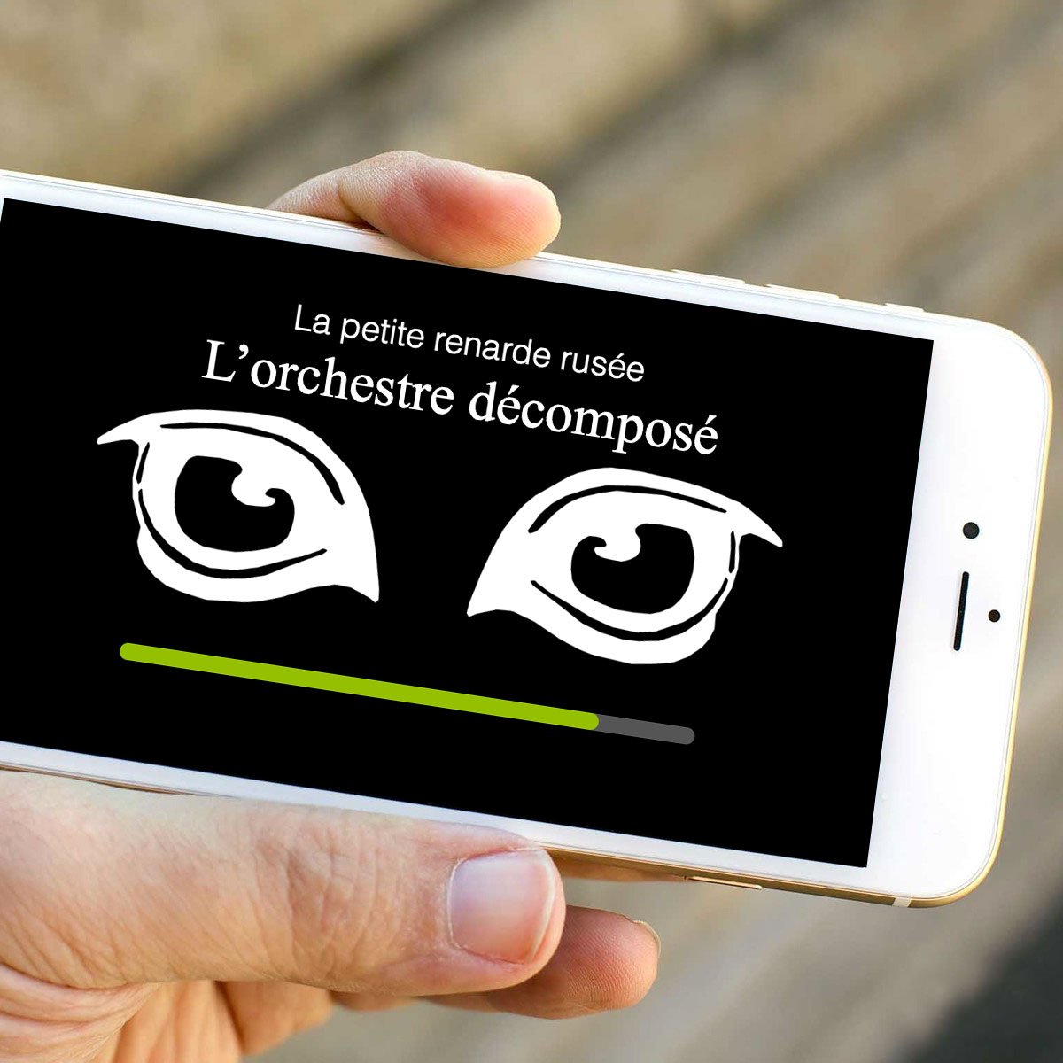 L'image montre un smartphone tenu dans une main. L'écran du téléphone affiche un fond noir avec deux yeux stylisés en blanc. Au-dessus des yeux, il y a un texte qui dit "La petite renarde rusée" et, en dessous, le titre "L'orchestre décomposé". Une barre de chargement verte est visible en bas de l'écran.