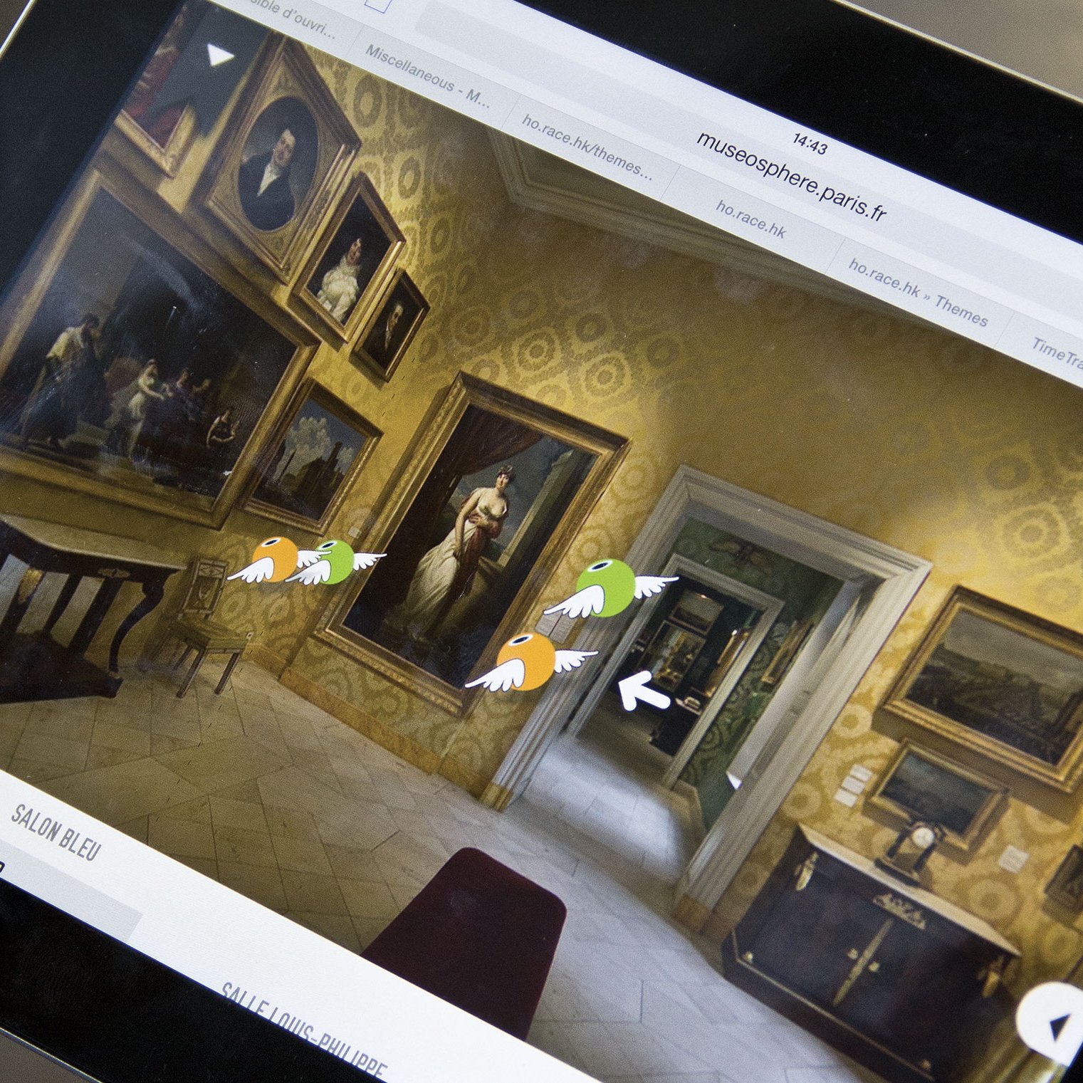 L'image montre une personne tenant une tablette affichant une visite virtuelle d'un musée. Sur l'écran, on peut voir une salle de musée avec des peintures accrochées aux murs, ainsi qu'un mobilier ancien. Les murs sont décorés de motifs et il y a des flèches indiquant les directions à explorer, suggérant une interface interactive pour naviguer dans l'espace.