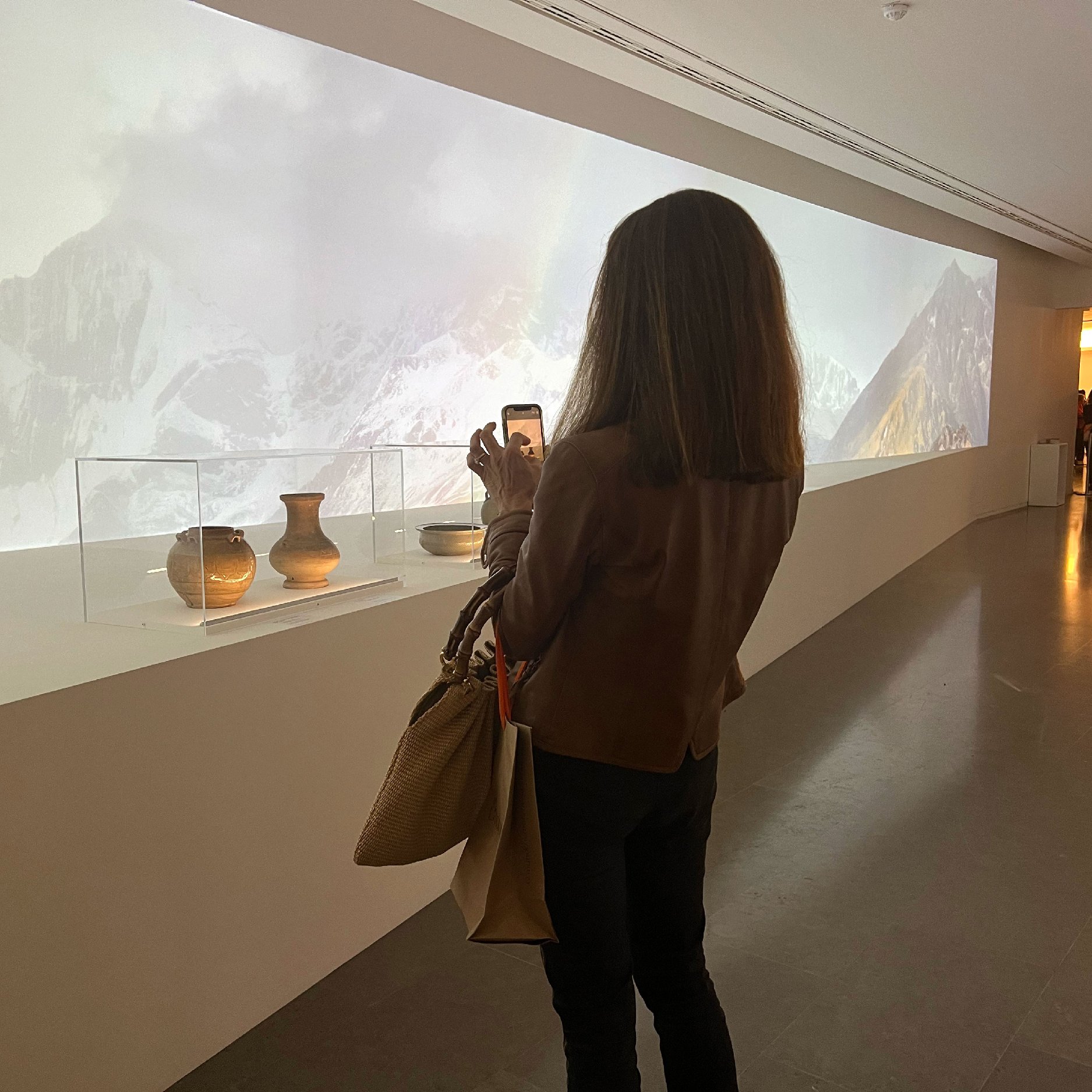 L'image montre une femme se tenant devant une exposition dans un musée. Elle est de dos, prenant une photo avec son téléphone. Devant elle, il y a une vitrine contenant plusieurs pots, tandis qu'en arrière-plan, on peut voir une projection de paysages de montagnes. L'éclairage est doux, et l'atmosphère semble calme et contemplative.