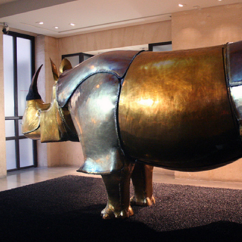 L'image montre une sculpture d'un rhinocéros, réalisée en métal poli. La sculpture a une finition dorée et argentée, ce qui lui donne un aspect brillant et artistique. Elle est placée dans un espace intérieur, probablement dans un musée ou une galerie d'art, avec un fond sobre et des lumières mettant en valeur les courbes de l'œuvre. Le rhinocéros se tient de profil, avec une posture imposante et élégante.