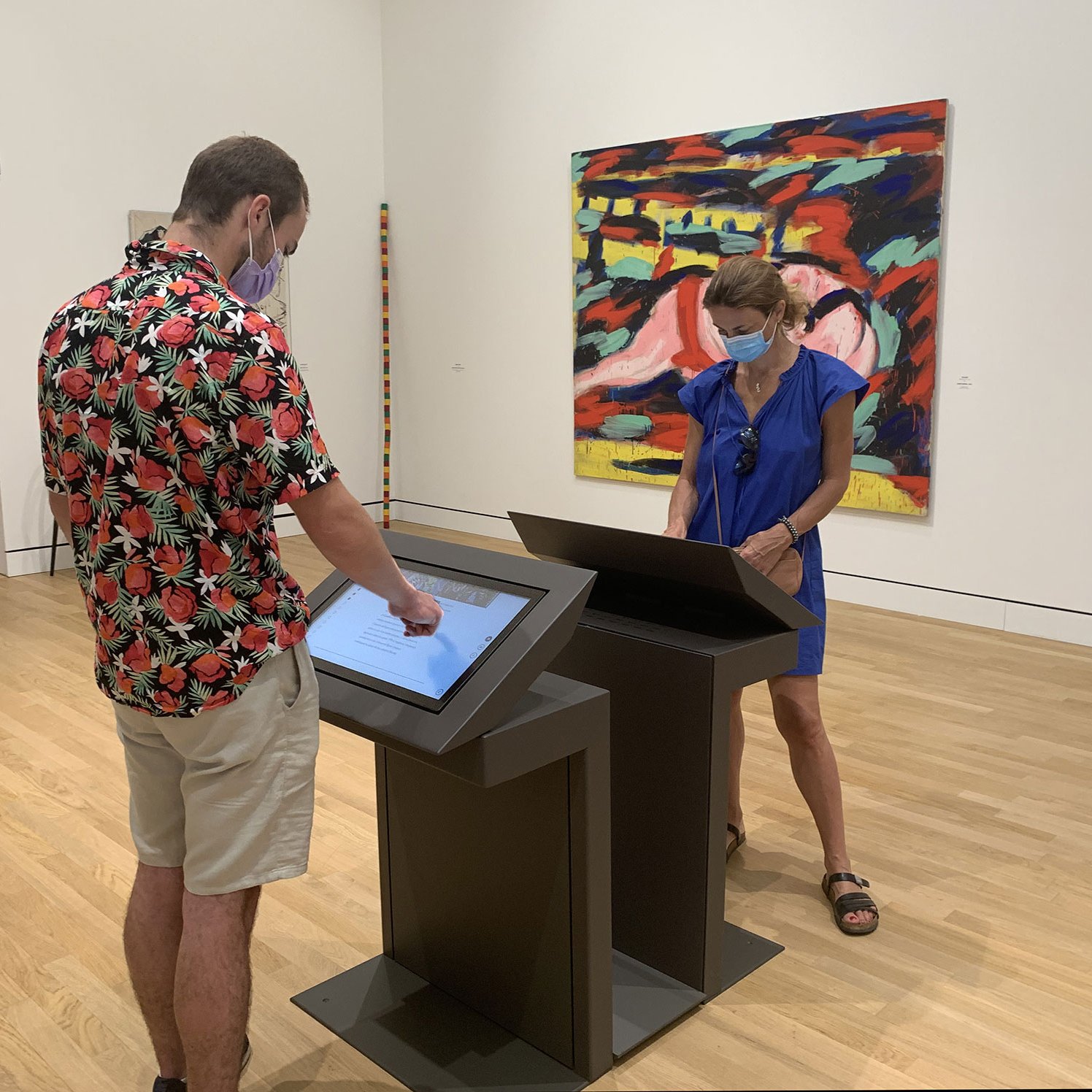 L'image montre deux personnes dans un musée ou une galerie d'art. L'une d'elles, un homme portant une chemise à motifs floraux, est en train d'interagir avec un écran tactile. L'autre personne, une femme en robe bleue, observe ce qu'il fait. Les deux portent des masques de protection. En arrière-plan, on peut voir une toile colorée accrochée au mur, ajoutant une ambiance artistique à l'espace. Le sol est en bois clair.