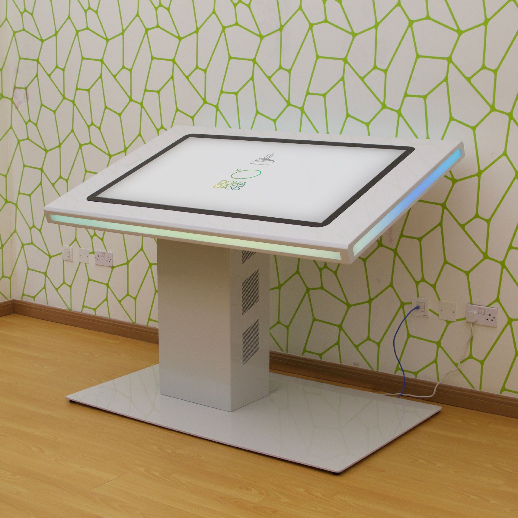 L'image montre une table interactive moderne qui semble conçue pour des utilisations technologiques ou éducatives. Elle est rectangulaire et inclinée, avec un écran tactile sur le dessus. La base de la table est en métal brillant, de couleur blanche, et elle repose sur un socle large. Le fond est orné d'un motif géométrique vert sur un mur clair, ajoutant une touche contemporaine à l'environnement. On peut également voir des prises électriques à proximité de la table.