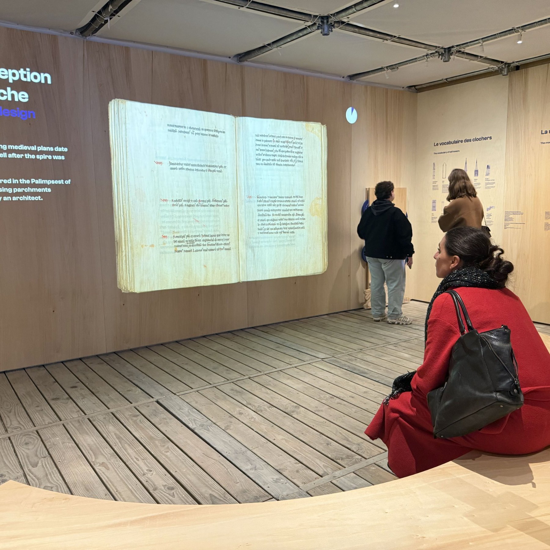 L'image montre une exposition dans un musée, où des visiteurs explorent des panneaux d'information. Au centre, on voit une femme assise sur une structure en bois, vêtue d'un manteau rouge et d'un sac à main noir. Les panneaux sur le mur incluent des informations sur le design des flèches architecturales, avec des illustrations et du texte en français. L'ambiance est moderne et épurée, avec des murs en bois et un sol en bois.