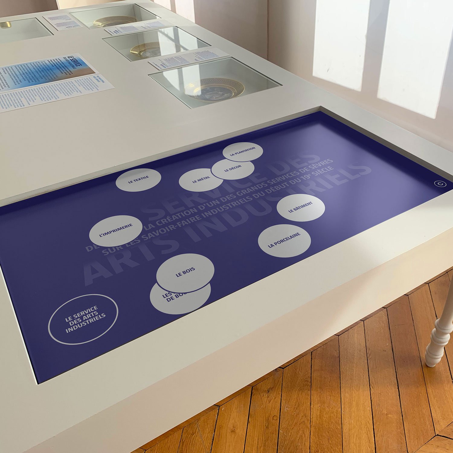 L'image montre une table d'exposition design moderne avec une surface interactive. La table semble être utilisée dans un contexte muséal ou éducatif. Elle possède des éléments ronds et des illustrations ou textes disposés autour d'un écran tactile au centre. Le fond de l'écran est de couleur violette et inclut des éléments graphiques liés aux arts industriels. La table a des pieds en bois, et le sol est en parquet. Les murs environnants semblent clairs, ce qui donne une atmosphère lumineuse à l'espace.