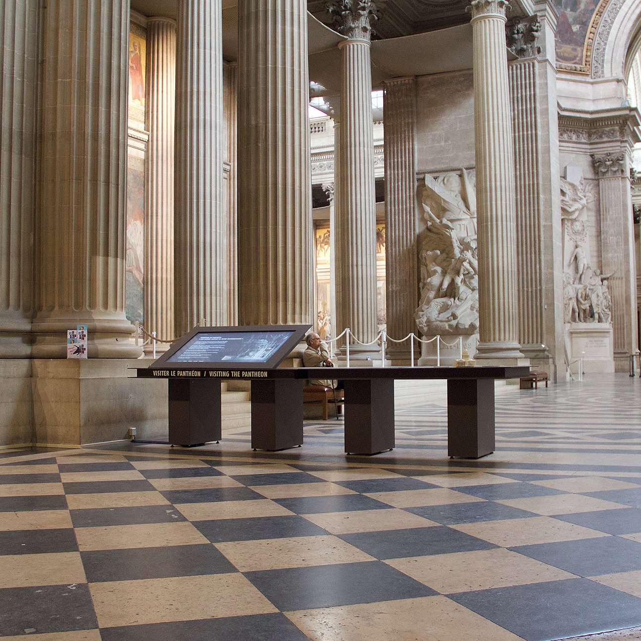 L'image montre l'intérieur d'un grand bâtiment avec des colonnes majestueuses et un sol en damier. On peut voir des sculptures et des fresques sur les murs, ajoutant à l'impression d'élégance et d'histoire de l'espace. Au centre, il y a une table interactive qui semble fournir des informations aux visiteurs. L'éclairage est doux, mettant en valeur les détails architecturaux. Des personnes se déplacent, ce qui indique que c'est un lieu ouvert au public.