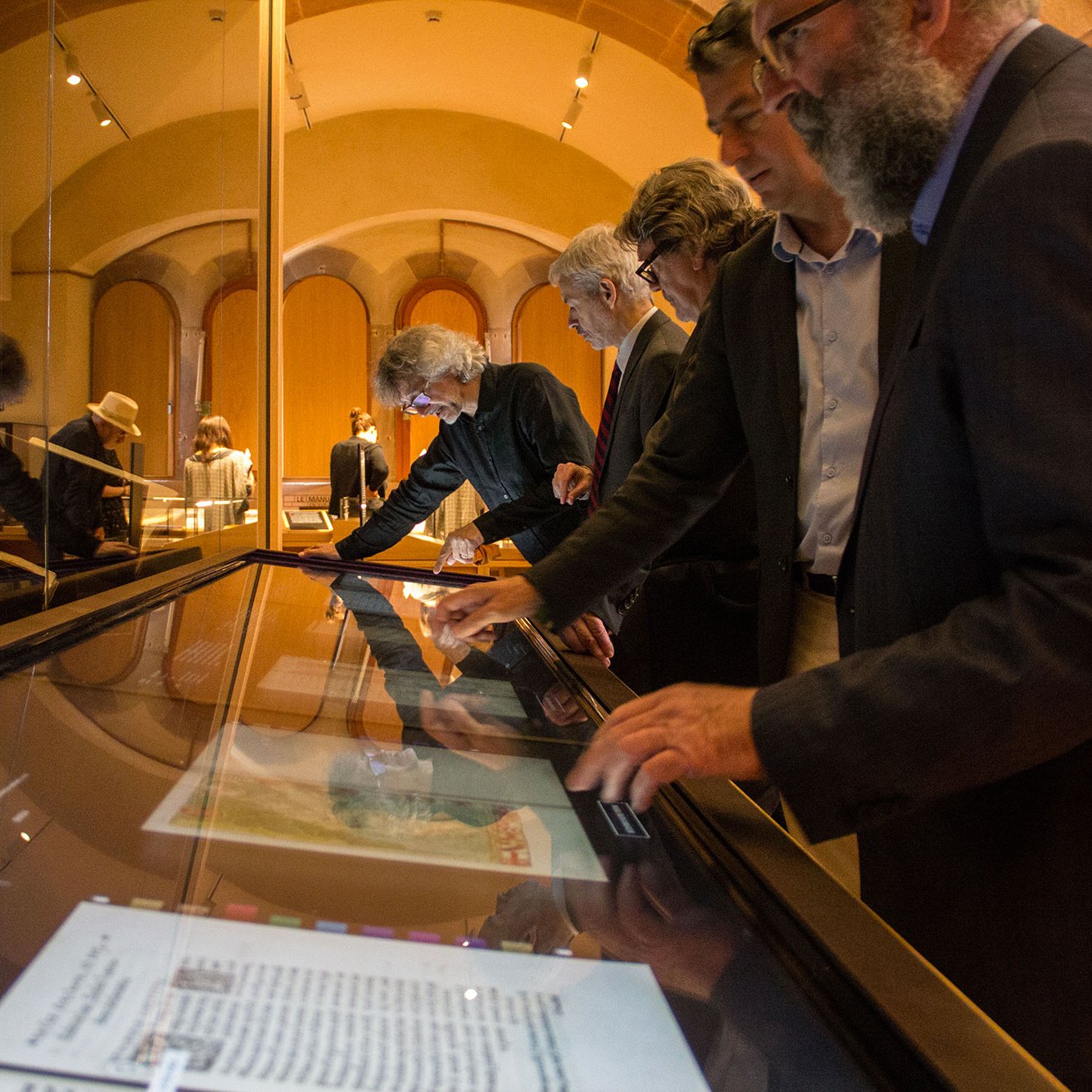 L'image montre un groupe de personnes debout devant une exposition dans un musée. Elles semblent intéressées et observent des objets présentés sur une table interactive. Les visiteurs, de différents âges et styles vestimentaires, touchent l'écran pour explorer les informations ou les images. En arrière-plan, on aperçoit des livres et des documents affichés dans des vitrines. L'éclairage est doux, créant une ambiance chaleureuse et propice à la découverte.