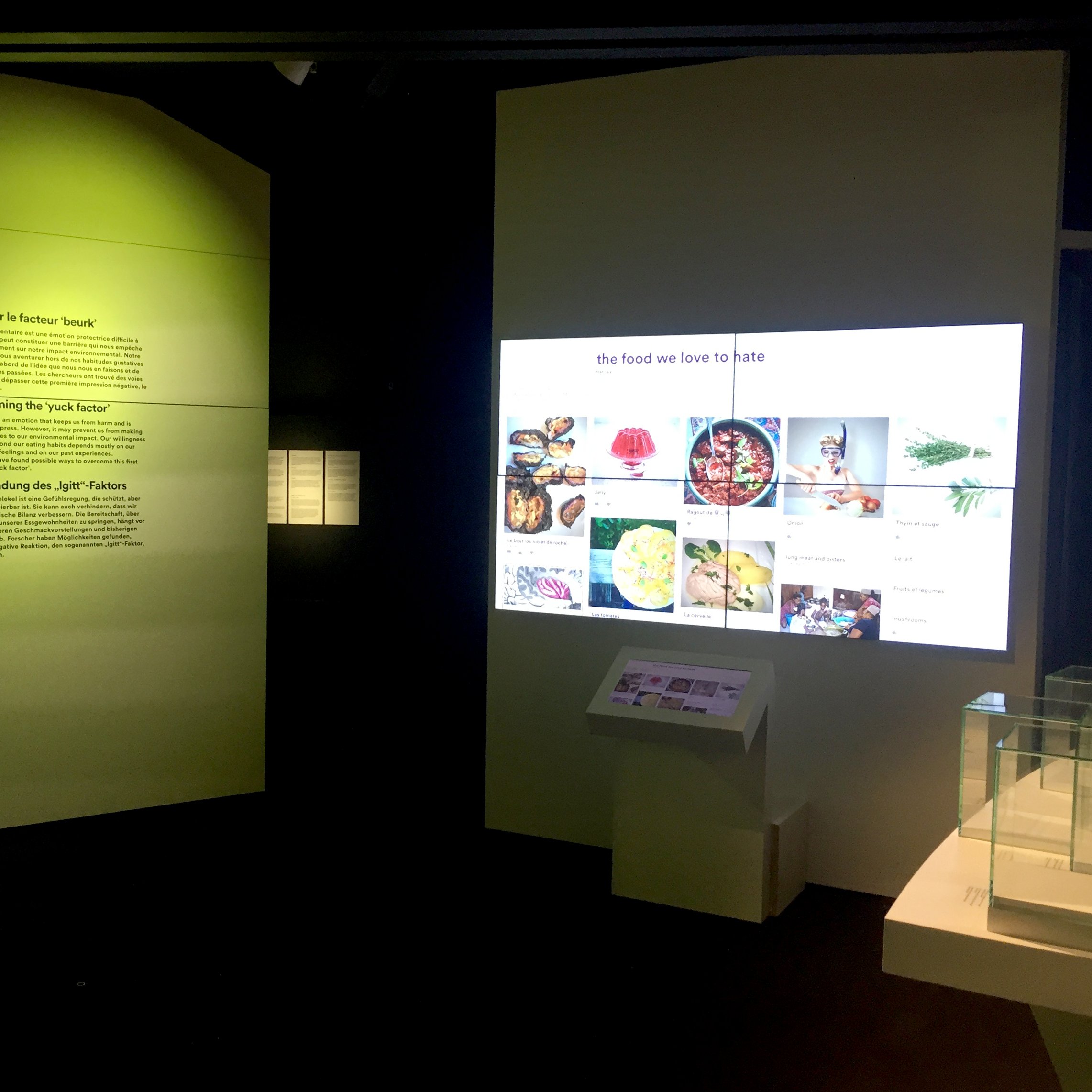 L'image montre une exposition dans un espace muséal ou culturel. À gauche, il y a un mur vert illuminé avec des textes écrits en français et en allemand, expliquant probablement un concept ou une thématique. À droite, un grand écran affiche des images de nourriture, suggérant une discussion sur les aliments que l'on peut aimer ou détester. Au bas de l'écran, un dispositif interactif semble présenter des informations supplémentaires. L'éclairage rend l'espace moderne et immersif.