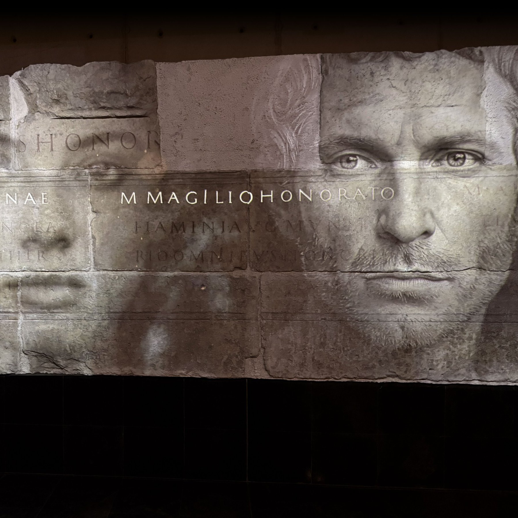 L'image présente une série de visages en noir et blanc, superposés de manière artistique sur une surface ressemblant à de la pierre ou du béton. Les traits des visages sont flous, créant une atmosphère mystérieuse. Au centre, des inscriptions en lettres capitales sont visibles, probablement des noms ou des titres. L'ensemble de la composition dégage une impression de gravité et d'histoire, suggérant une connexion avec des thèmes de mémoire ou d'honneur.