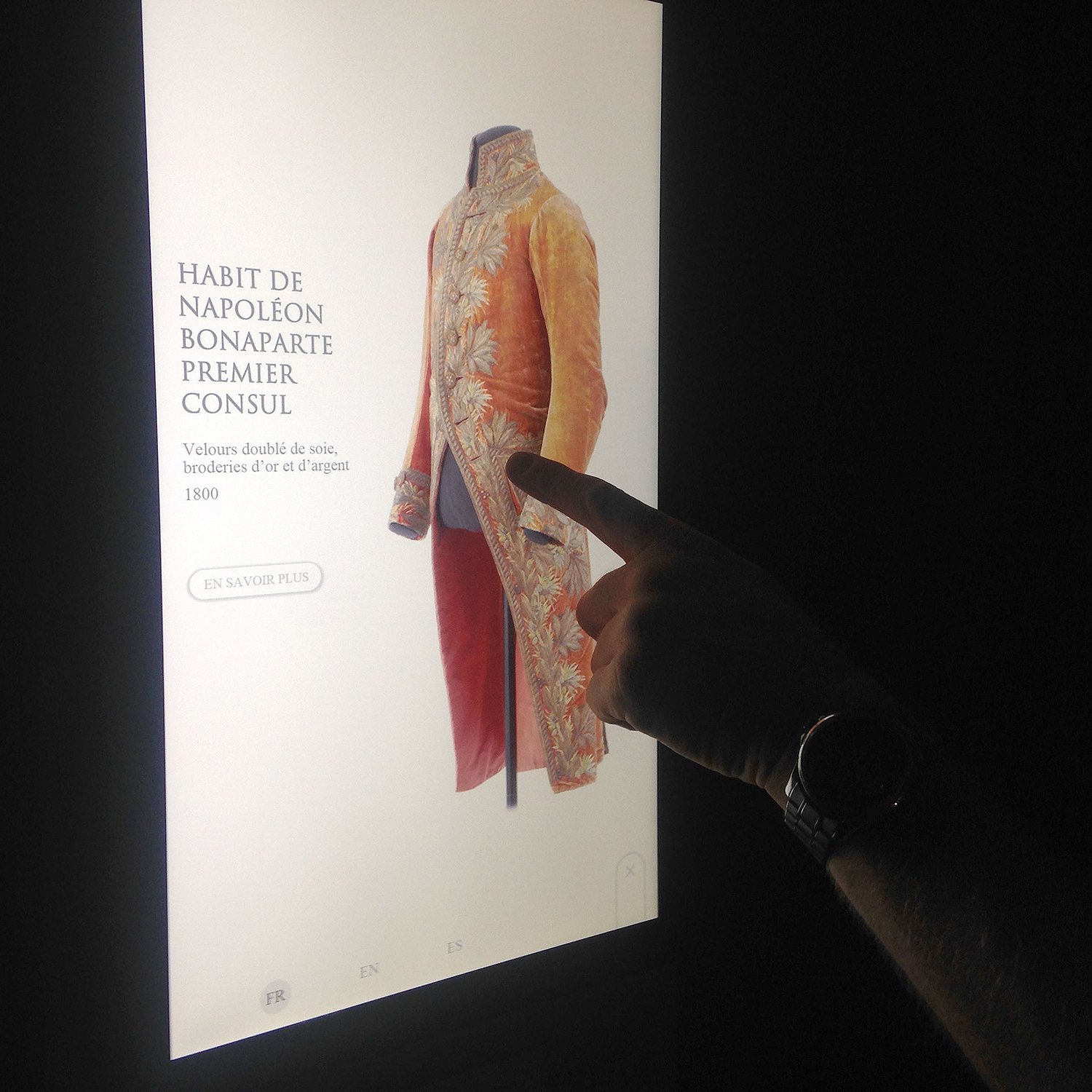 L'image montre un écran interactif présentant un habit de Napoléon Bonaparte, qui était Premier Consul. Le vêtement est richement décoré, en soie, avec des broderies. Une main se tient près de l'écran, soulignant le costume, et il y a une option pour en savoir plus en bas de l'image.