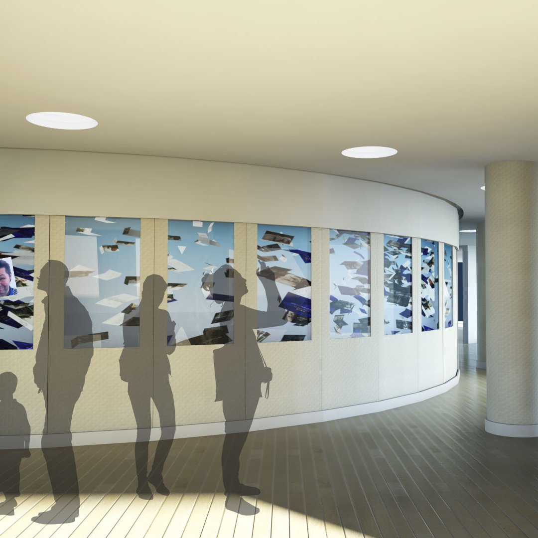 L'image montre un intérieur moderne avec un espace circulaire. Sur un mur, il y a une exposition de photos ou d'œuvres artistiques présentées dans des cadres. Au premier plan, on aperçoit des silhouettes de personnes, dont un enfant. L'éclairage est doux, et le sol est en bois, créant une ambiance chaleureuse. À l'arrière-plan, on distingue un escalier moderne qui mène à un autre niveau. L'ensemble de la scène semble contemporain et accueillant.