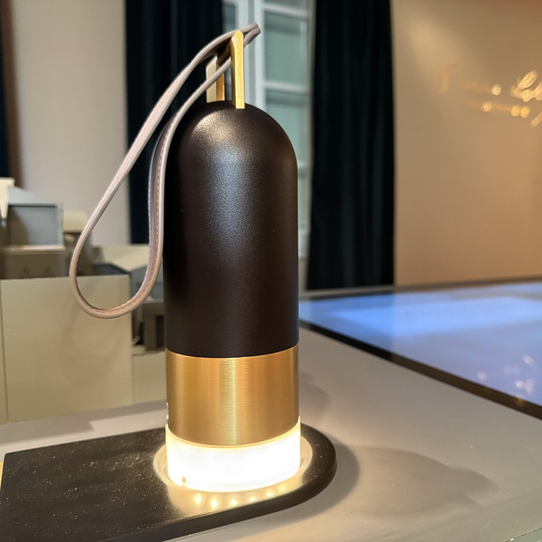 L'image montre un objet qui ressemble à une lampe ou un éclairage de table. Il a une forme cylindrique avec une partie supérieure en noir et une base en métal doré. Un cordon est attaché à la partie supérieure, ce qui suggère qu'il peut être déplacé facilement. La base est illuminée par une lumière douce, ajoutant une ambiance chaleureuse. L'objet est placé sur une surface moderne et épurée, avec un arrière-plan flou.