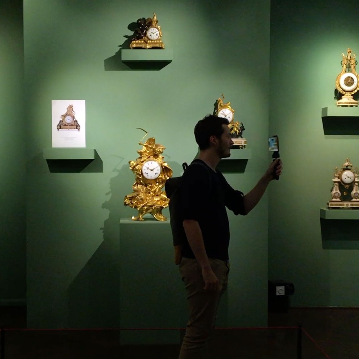 L'image montre une exposition dans un musée ou une galerie. Sur le mur vert, plusieurs horloges décoratives sont présentées sur des étagères. L'une des horloges est encadrée par une plaque explicative. Au premier plan, un homme tient un téléphone, probablement pour prendre des photos ou enregistrer des informations. L'éclairage met en valeur les détails des horloges, qui semblent être des pièces artistiques anciennes et ornées.