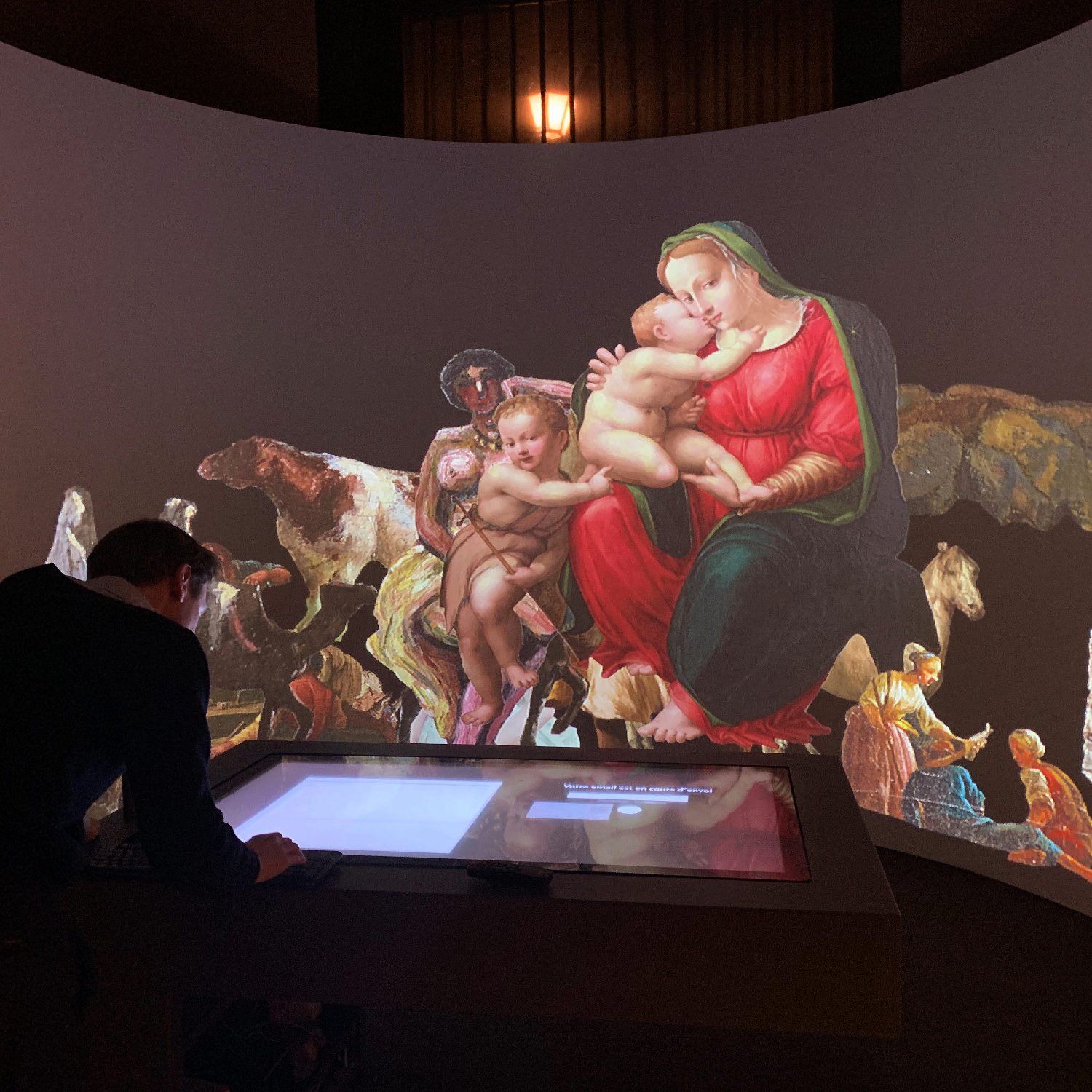 L'image montre une installation artistique immersive où une grande reproduction d'une œuvre d'art classique est projetée sur un mur. Un homme est penché sur une table interactive, semblant interagir avec l'exposition. En arrière-plan, des figures et des scènes colorées sont affichées, créant une atmosphère dynamique. Les éléments de l'œuvre incluent une Vierge à l'Enfant entourée de diverses figures humaines et animales, le tout dans un contexte artistique riche et captivant.