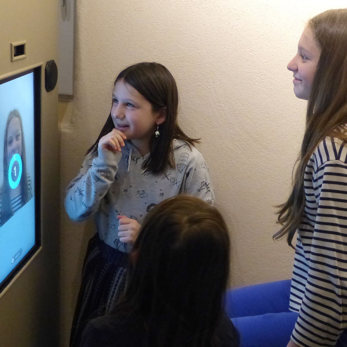 L'image montre un groupe de jeunes filles interagissant avec un écran. L'une d'elles, qui se tient devant l'écran, semble surprise ou amusée, tandis que les autres regardent avec intérêt. L'écran affiche une image qui pourrait être un reflet ou une vidéo d'elles. L'ambiance semble conviviale et ludique. Les filles portent des vêtements décontractés, ce qui renforce l'impression d'un moment informel et joyeux.