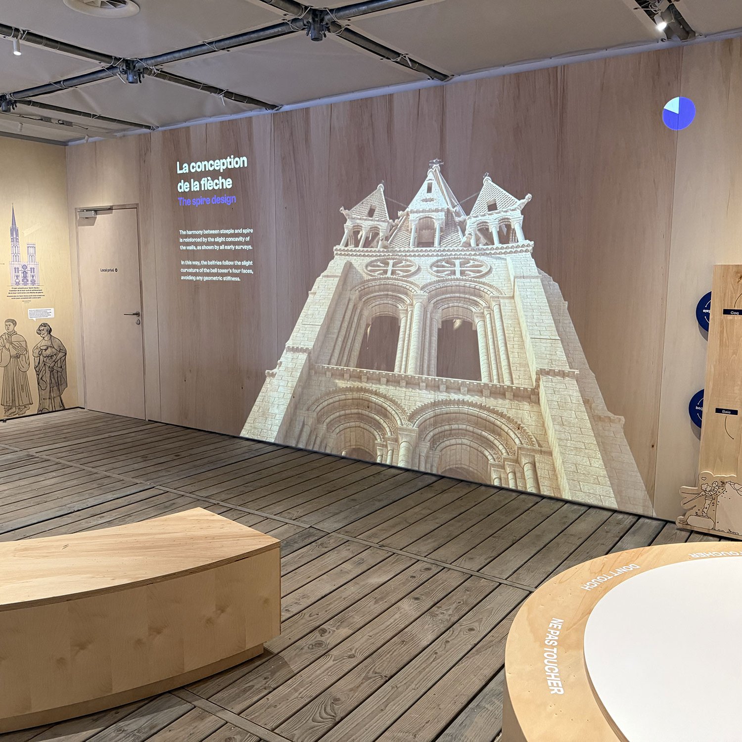 L'image montre un espace intérieur, probablement une exposition. On y voit un mur en bois et une projection d'une façade d'église, mettant en avant des éléments architecturaux comme des arcs et des fenêtres. À côté, il y a des illustrations et des textes explicatifs sur la conception d'une flèche. Le sol semble en bois, et il y a des structures en forme de demi-cercle qui pourraient servir de sièges ou de supports d'exposition. L'ensemble donne une impression moderne et éducative, mêlant art et histoire.