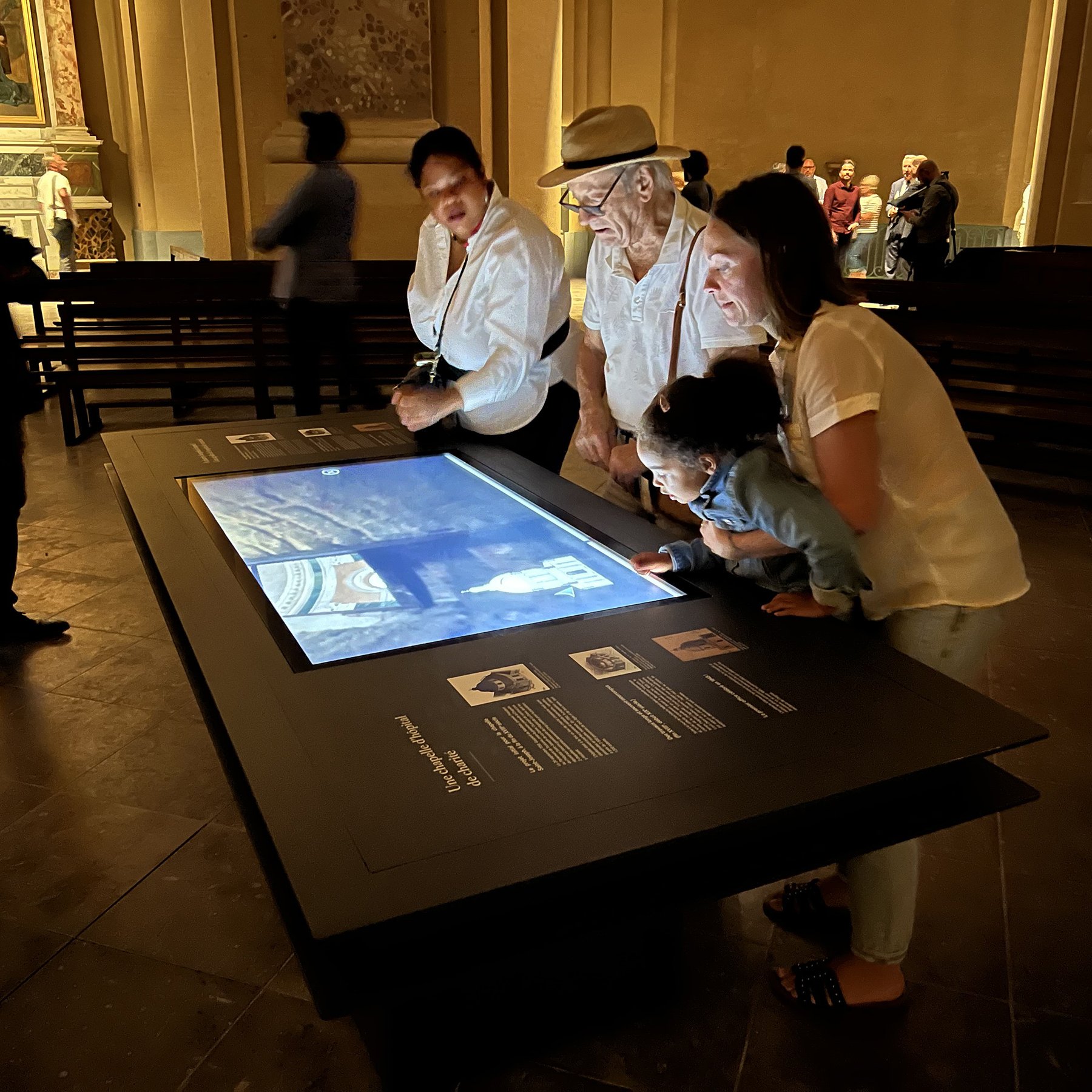 L'image montre un groupe de quatre personnes en train d'observer une table interactive au musée ou dans un espace d'exposition. On peut voir deux adultes et un enfant, concentrés sur l'écran qui affiche probablement des informations ou des images. Une ambiance de curiosité et d'engagement se dégage, alors que d'autres visiteurs se déplacent en arrière-plan. L'éclairage et le décor semblent suggérer un lieu culturel ou historique.