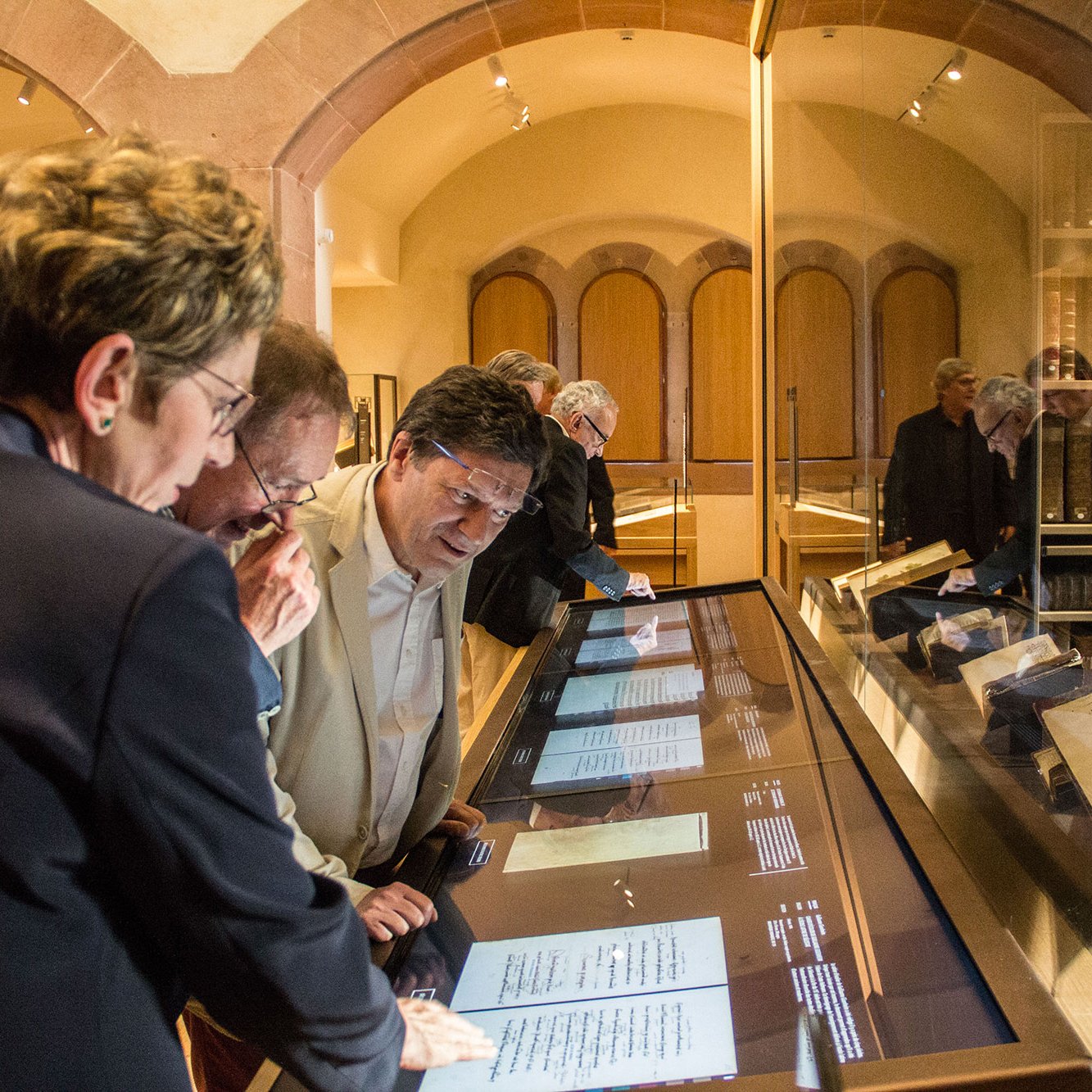 L'image montre un groupe de personnes réunies autour d'une vitrine dans un musée. Ils examinent des documents ou des manuscrits exposés, probablement de grande valeur historique. Les visiteurs semblent intéressés et engagés dans leur découverte, avec des expressions de curiosité. La lumière de l'espace muséal met en avant les objets exposés, créant une atmosphère d'exploration et d'apprentissage. En arrière-plan, d'autres personnes floues sont également présentes, profitant de l'exposition.