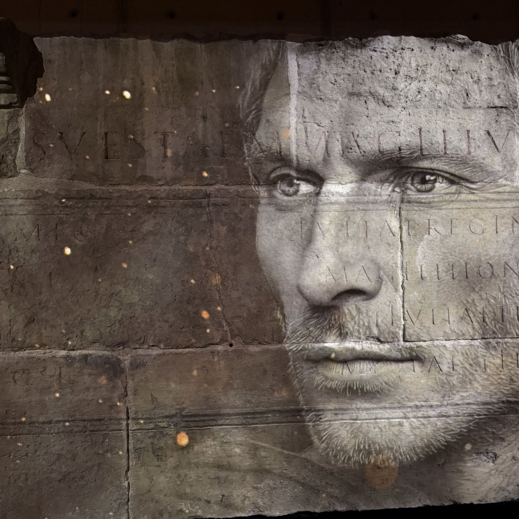 L'image présente une série de visages en noir et blanc, superposés de manière artistique sur une surface ressemblant à de la pierre ou du béton. Les traits des visages sont flous, créant une atmosphère mystérieuse. Au centre, des inscriptions en lettres capitales sont visibles, probablement des noms ou des titres. L'ensemble de la composition dégage une impression de gravité et d'histoire, suggérant une connexion avec des thèmes de mémoire ou d'honneur.