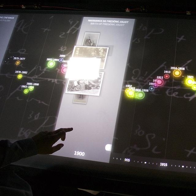 L'image montre une interface interactive, probablement un écran tactile ou un tableau numérique, présentant une chronologie historique. À gauche, on peut voir des dates allant de 1900 à environ 1915, accompagnées de photographies anciennes. Des cercles colorés représentent des événements marquants, montrant l'évolution sur une période donnée. Des éléments graphiques, tels que des notes manuscrites, ajoutent un aspect visuel intéressant à l'ensemble. Un doigt humain pointe vers l'écran, suggérant une interaction avec l'information présentée.