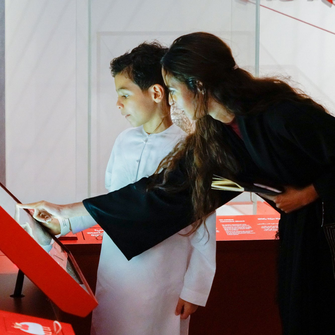 The image shows a young boy and a woman interacting with a display at an exhibition. The boy, wearing a white traditional outfit, is closely observing the screen while the woman, dressed in a dark garment, points to something on the display. Both appear engaged and curious as they explore the exhibit. The environment is modern, with red and white colors dominating the design.