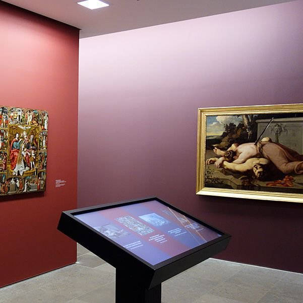 L'image montre une exposition dans un musée. Sur un mur d'une teinte rougeâtre, il y a un texte en blanc qui indique "ÉTUDES PRÉLIMINAIRES : LE CONSTAT D'ÉTAT". À côté, on peut voir une œuvre artistique encadrée, représentant une scène avec des figures humaines. Au premier plan, il y a un écran ou une table interactive qui semble contenir plus d'informations ou des illustrations liées à l'exposition. L'espace semble bien éclairé et organise une mise en valeur des œuvres présentées.