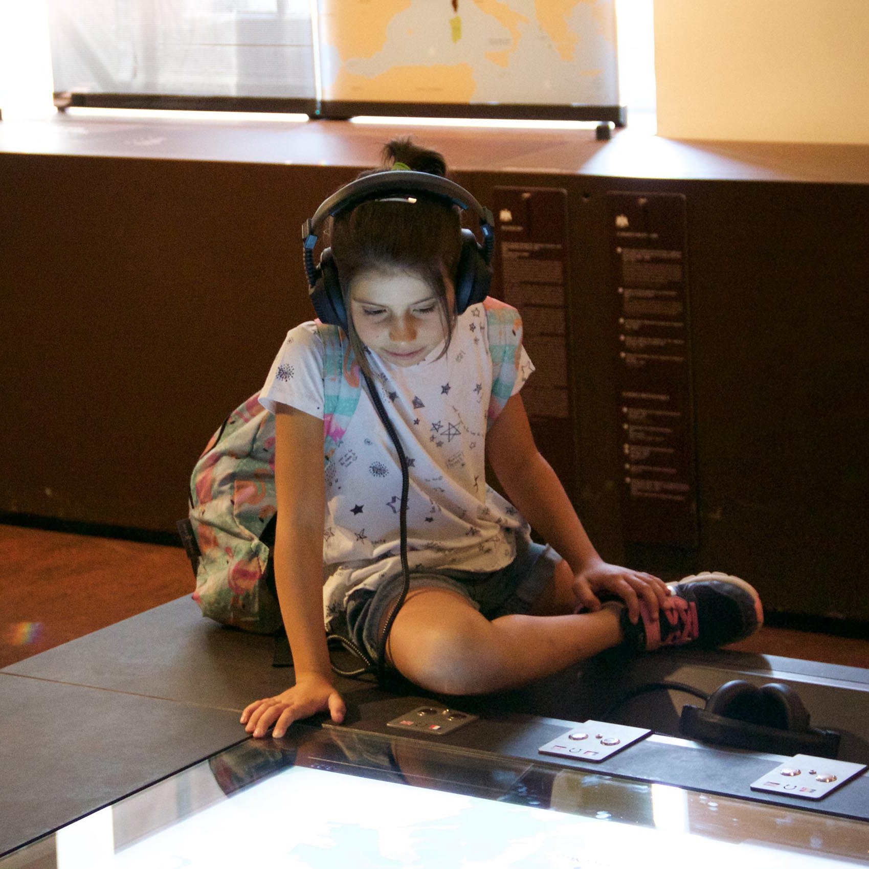 The image shows a young girl sitting on the floor at a museum or interactive exhibit. She is wearing headphones and appears to be focused on a touchscreen display in front of her. The girl has a backpack beside her and is dressed in casual clothing, including a T-shirt with a star pattern and shorts. The atmosphere looks educational, with various informational panels in the background.