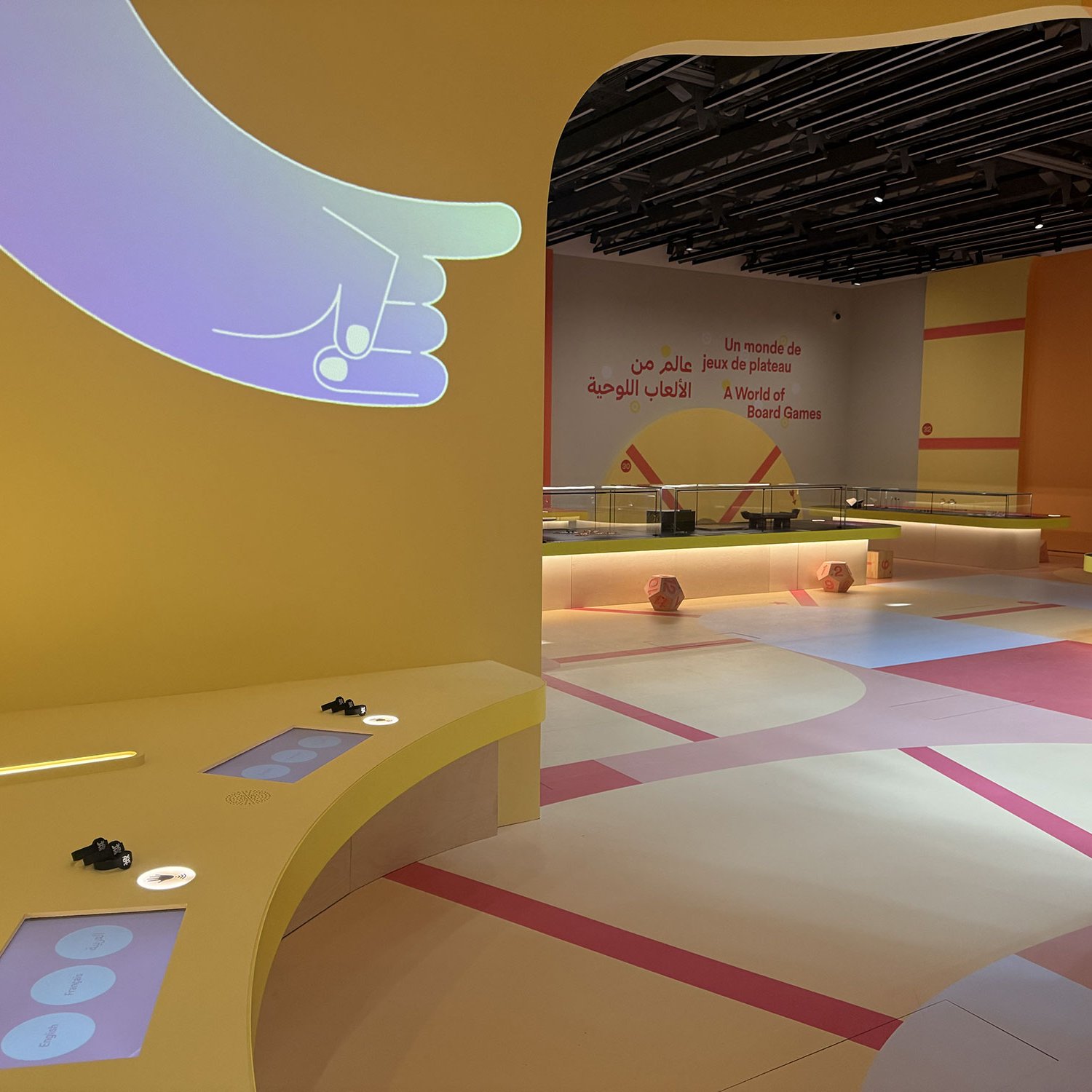 L'image montre un espace intérieur coloré avec des murs jaunes et des éléments de design modernes. On peut voir des tables avec des dispositifs interactifs et de l'éclairage doux. À l'arrière-plan, il y a des inscriptions en arabe et en français, indiquant une exposition sur les jeux de société. L'agencement de la pièce semble accueillant et ludique, avec des tapis de sol aux couleurs variées, créant une atmosphère conviviale.