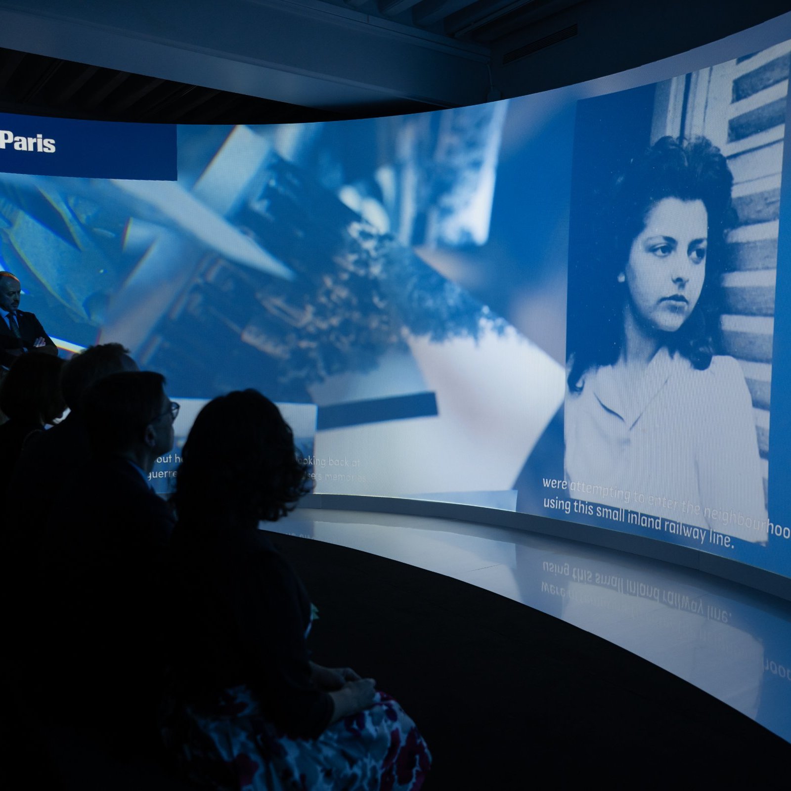 L'image montre une installation artistique ou un événement avec un écran incurvé affichant des contenus visuels. À gauche, une personne semble présenter ou parler, tandis qu'à droite, on voit une photo en noir et blanc d'une femme. Le fond est illuminé et les spectateurs, assis en face, regardent attentivement. Le mot « Paris » est affiché en haut. L'atmosphère semble être celle d'une exposition ou d'une présentation multimédia.