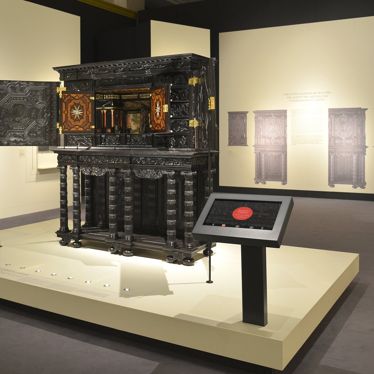 L'image montre une exposition muséale où un meuble ou une armoire ornée est mise en valeur au centre. Ce meuble, riche en détails, possède des sculptures et des marqueteries colorées. Des panneaux d'information sont positionnés près de l'objet, expliquant probablement son histoire ou son origine. À l'arrière-plan, on aperçoit un tableau dans un style classique, ainsi que des reproductions ou des œuvres similaires sur les murs. L'éclairage de l'espace met en avant les détails du meuble, créant une atmosphère d'admiration pour cet objet d'art.