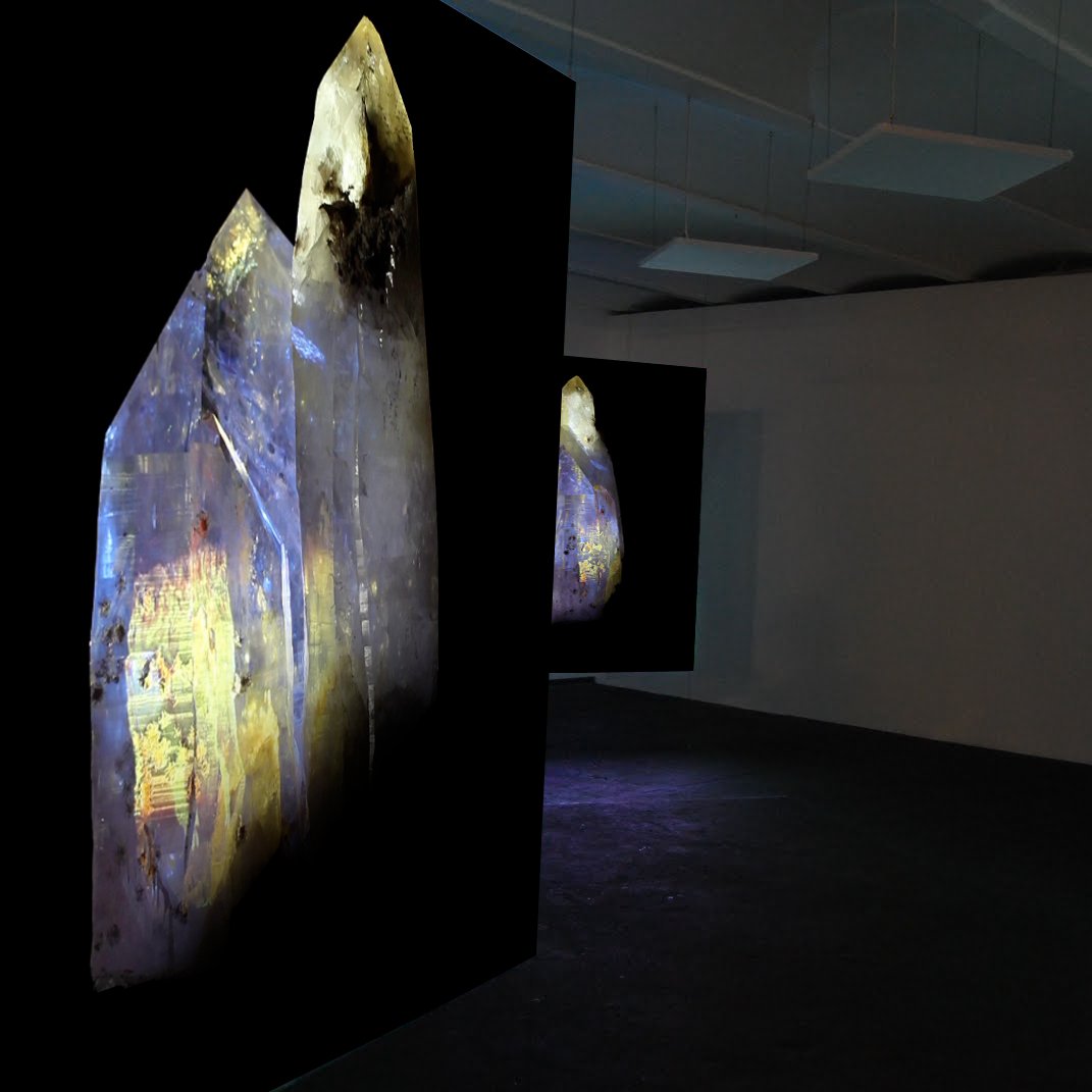 L'image montre une installation artistique dans un espace intérieur, probablement une galerie d'art. Plusieurs grands panneaux noirs sont suspendus au plafond, chacun affichant des images de formes rocheuses ou cristallines. Les couleurs sur ces panneaux varient, incluant des nuances de jaune, bleu et violet, ce qui crée un contraste intéressant avec le fond sombre. L'éclairage semble tamisé, mettant en valeur les détails des images et suggérant une atmosphère contemplative.