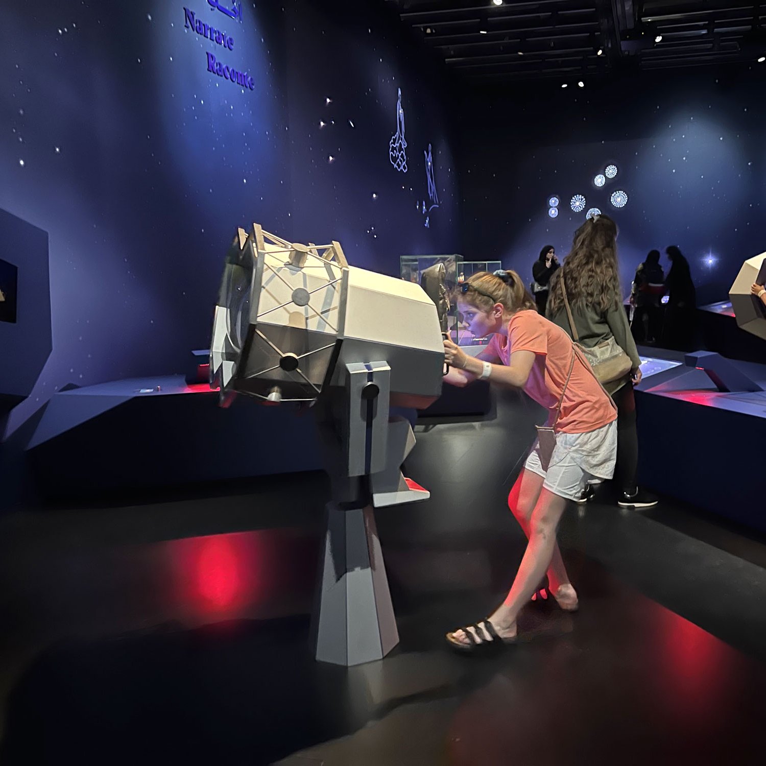 L'image montre une exposition dans un musée ou un espace éducatif. Une jeune fille se penche sur un appareil interactif ou une installation en forme de cylindre. Elle semble curieuse, examinant l'objet de près. En arrière-plan, plusieurs visiteurs explorent l'exposition, tandis que des éléments d'affichage et des décorations murales évoquent des thèmes scientifiques ou astronomiques. L'éclairage est doux, créant une ambiance agréable.