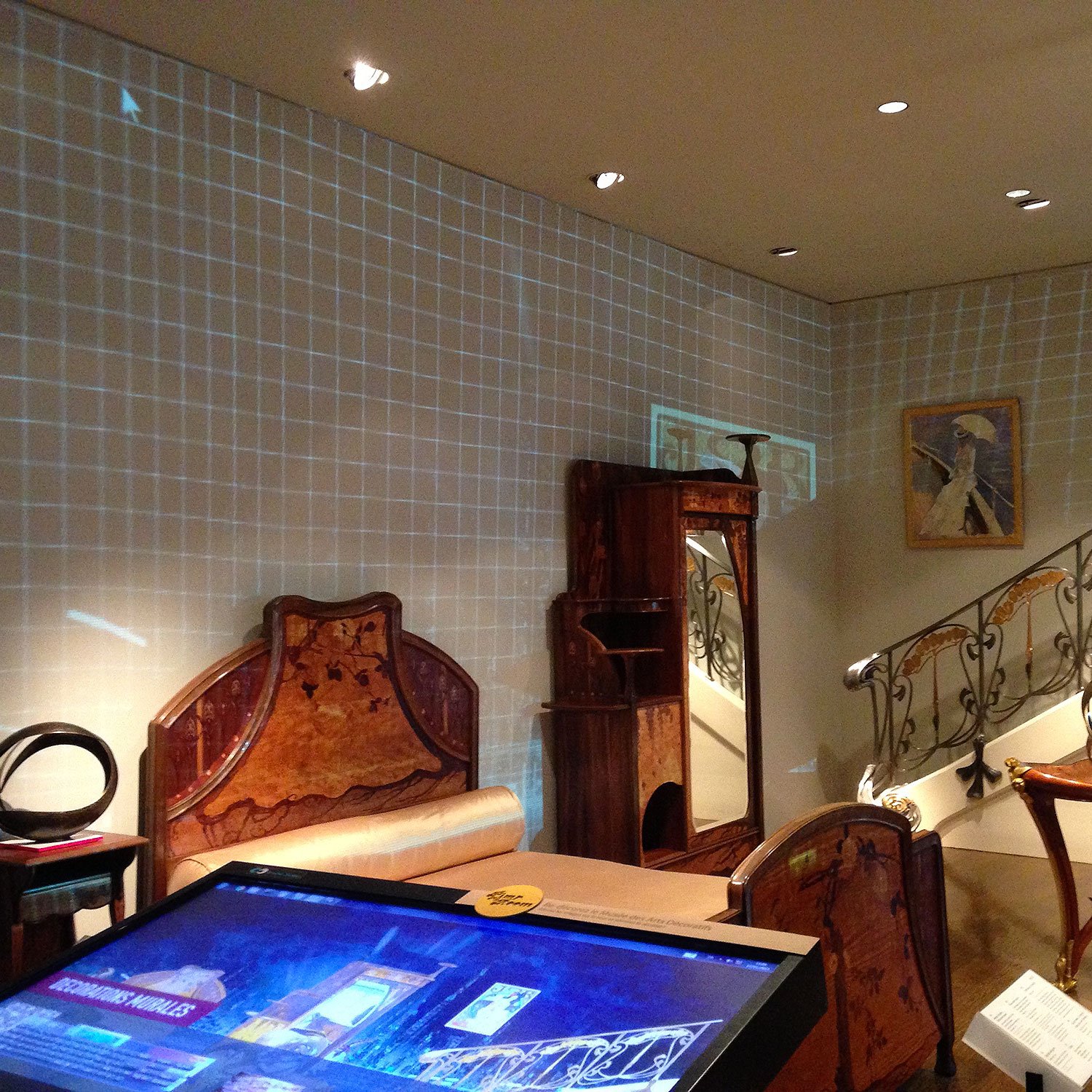 L'image montre une pièce aménagée de manière élégante, avec des meubles en bois sculpté. On peut voir un canapé et une chaise ornés, ainsi qu'un paravent. Une décoration murale projetée crée un effet de grille sur le mur, ajoutant une touche moderne à l'ensemble. Un escalier avec une rampe décorative se trouve à l'arrière-plan, tandis qu'un écran interactif est placé au premier plan, probablement utilisé pour fournir des informations sur les objets exposés. L'éclairage est doux et met en valeur le design des meubles.